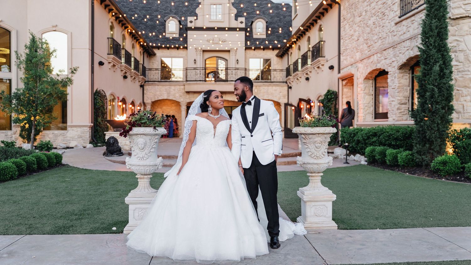 A romantic wedding couple strolls through an elegant courtyard with stone columns and lush greenery at dusk.