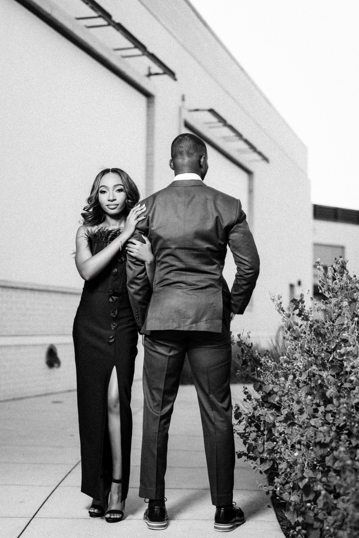 A couple shares a playful moment together against a white exterior wall with greenery.
