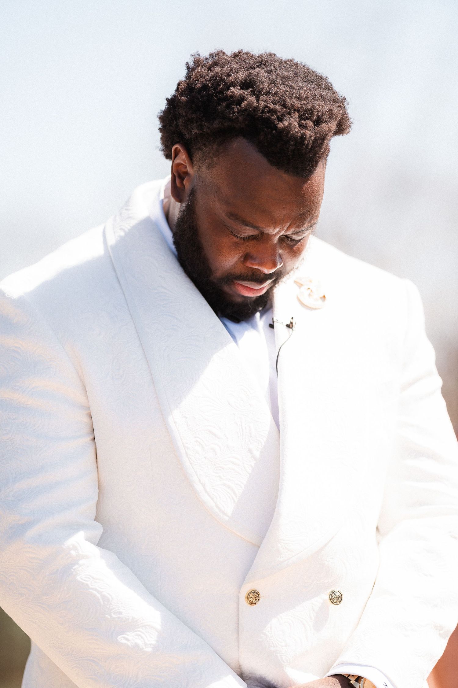 A person in a crisp white tuxedo jacket looks down thoughtfully in bright sunlight.