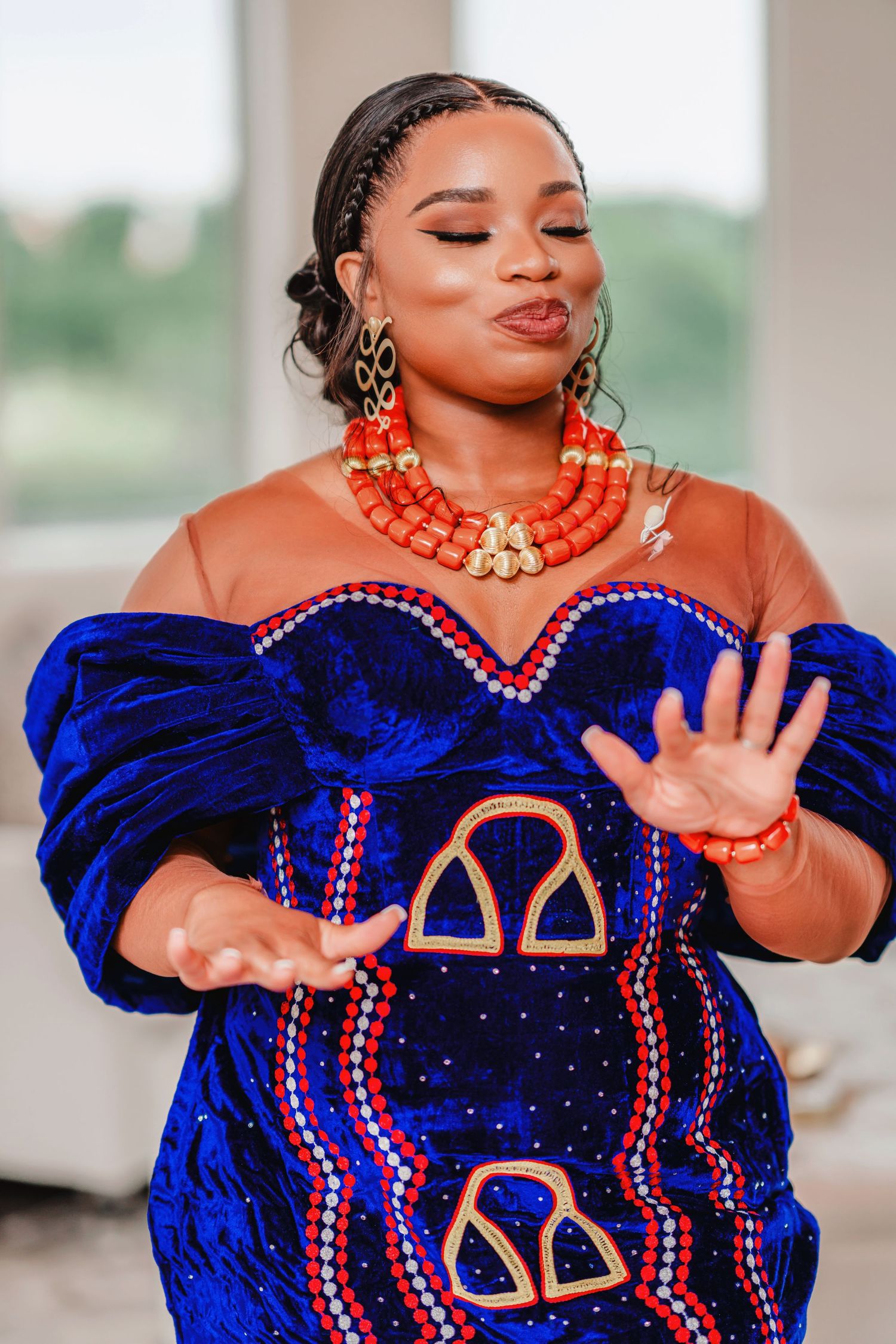 A woman in a royal blue velvet dress with gold accents and coral jewelry poses elegantly.