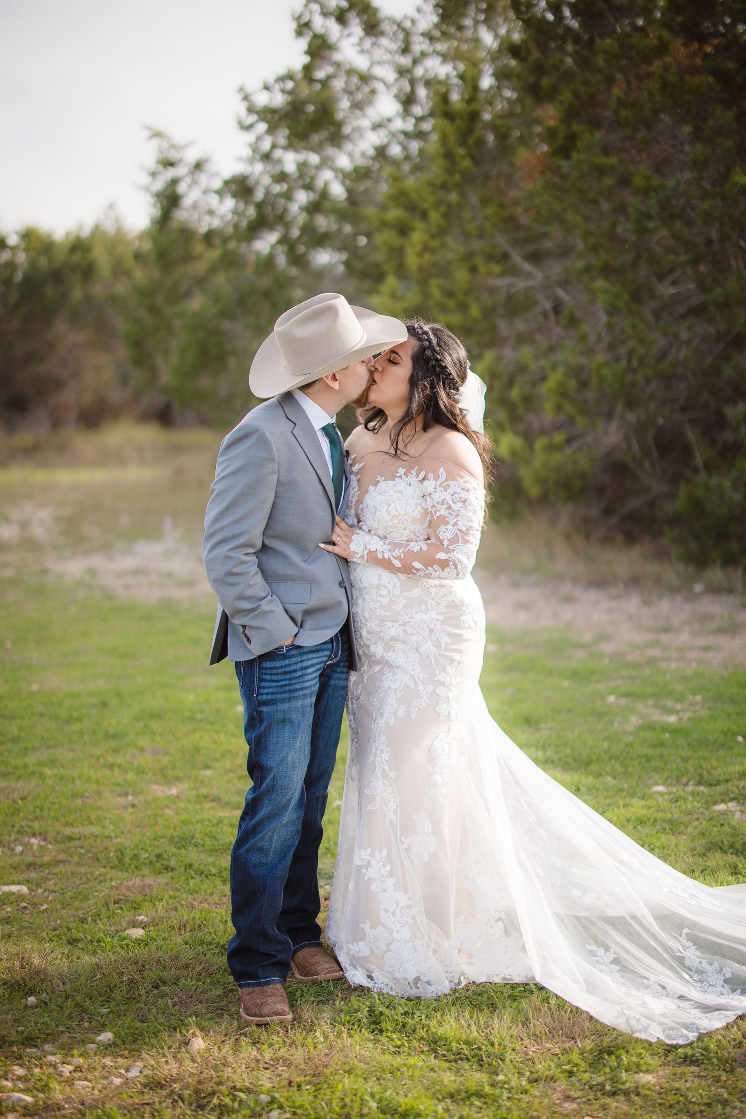 A romantic wedding couple embraces outdoors, with the bride in a lace gown and groom wearing a cowboy hat and gray jacket.