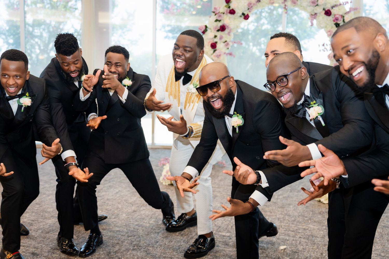 Wedding party members in black suits strike playful poses in a line against a light background.