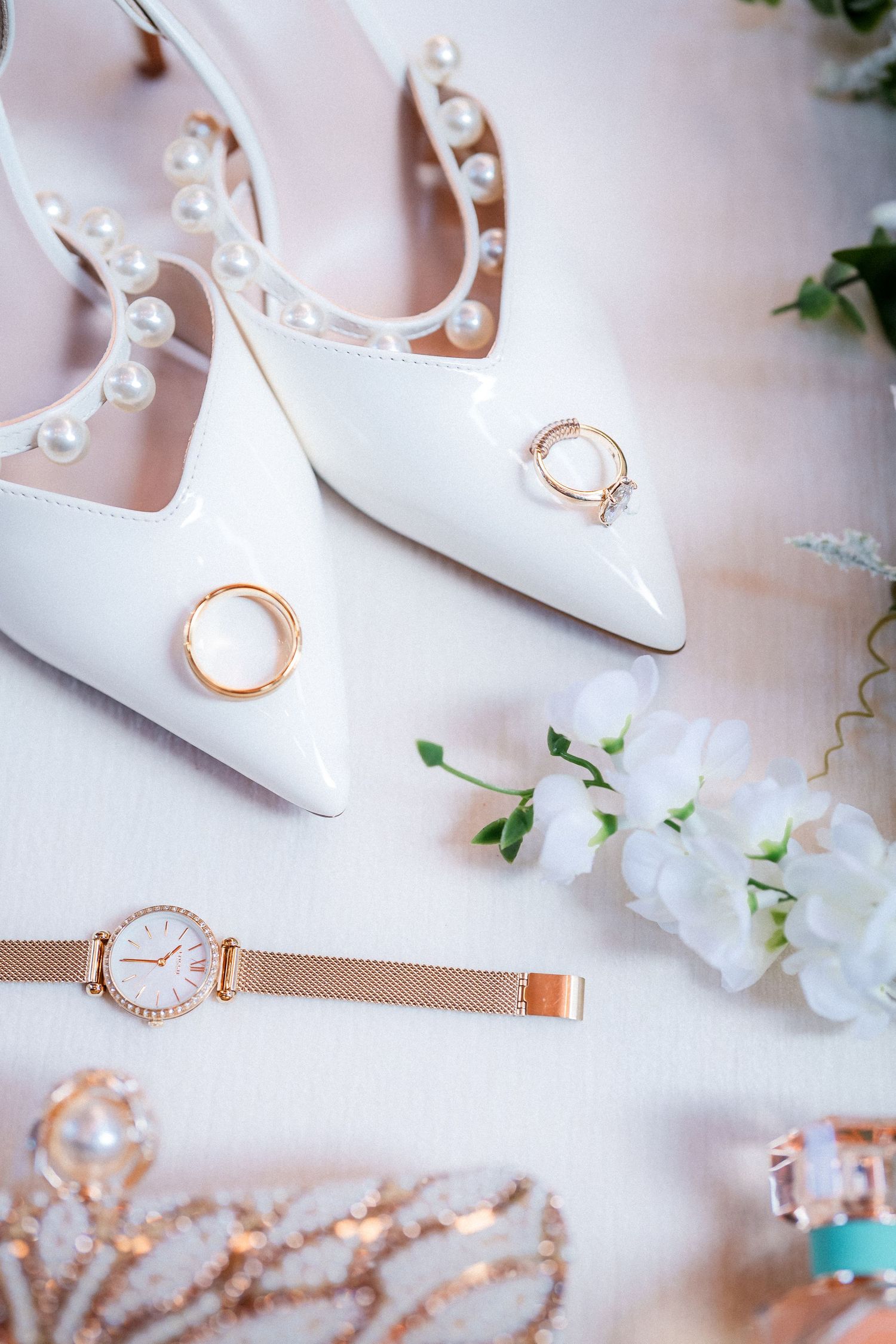 Wedding accessories including white heels, rings, and a rose gold watch arranged on a light surface.