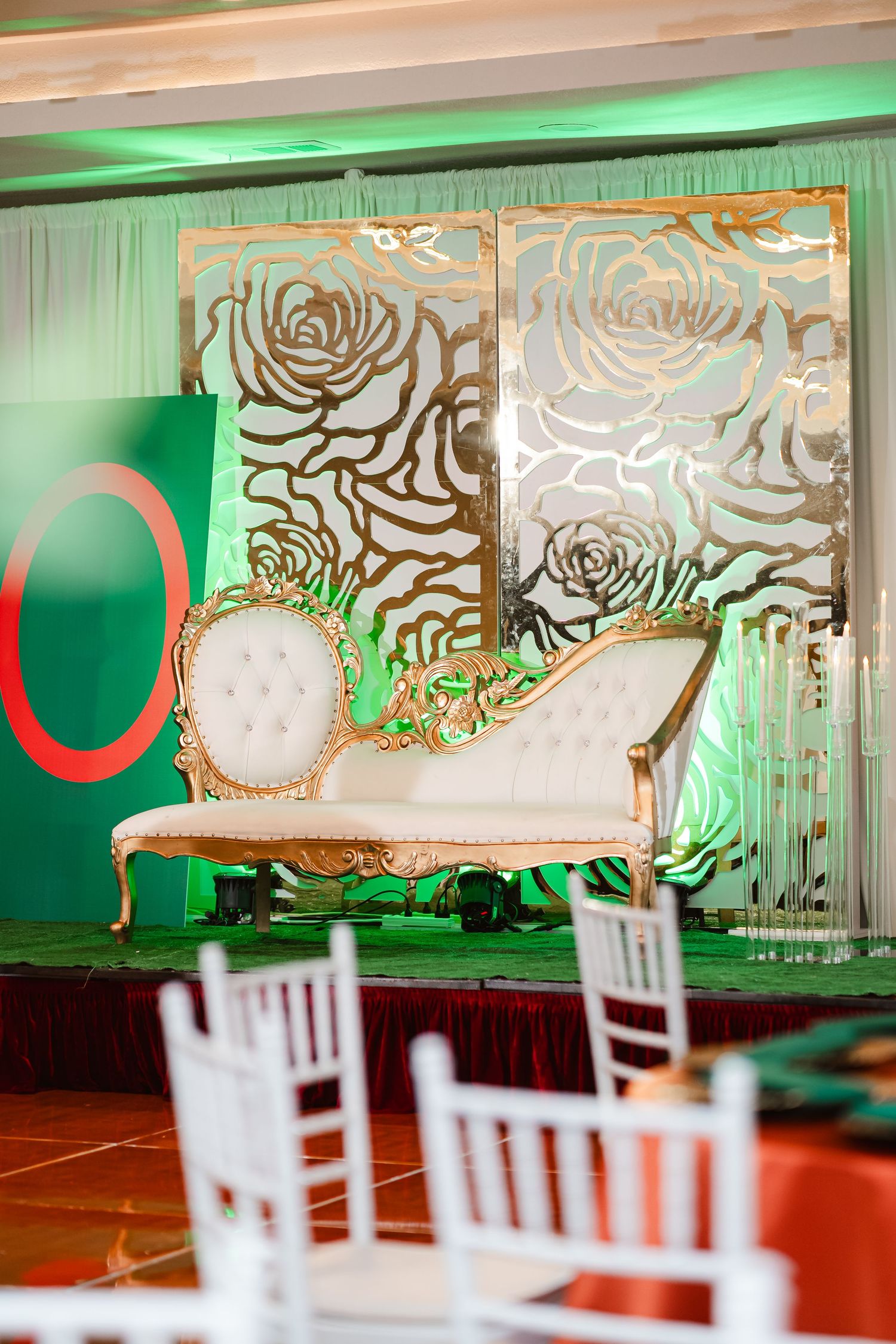 Elegant wedding reception venue with ornate gold and white backdrops illuminated by green uplighting and white chairs.
