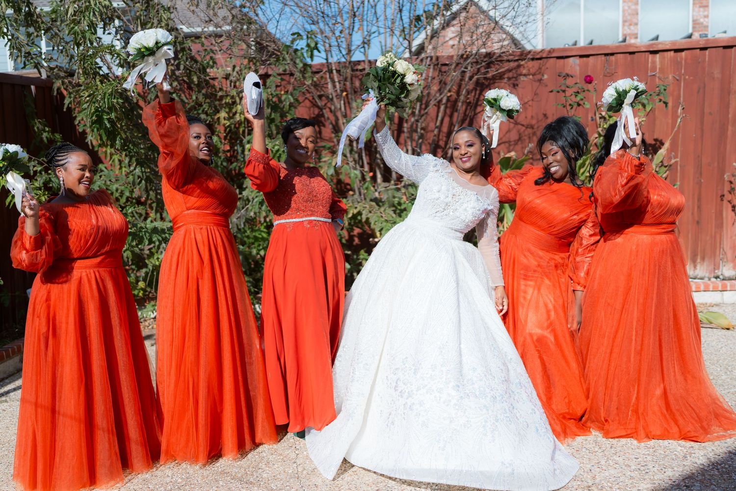 A bridal party celebrates outdoors wearing vibrant orange floor-length dresses with white floral bouquets.