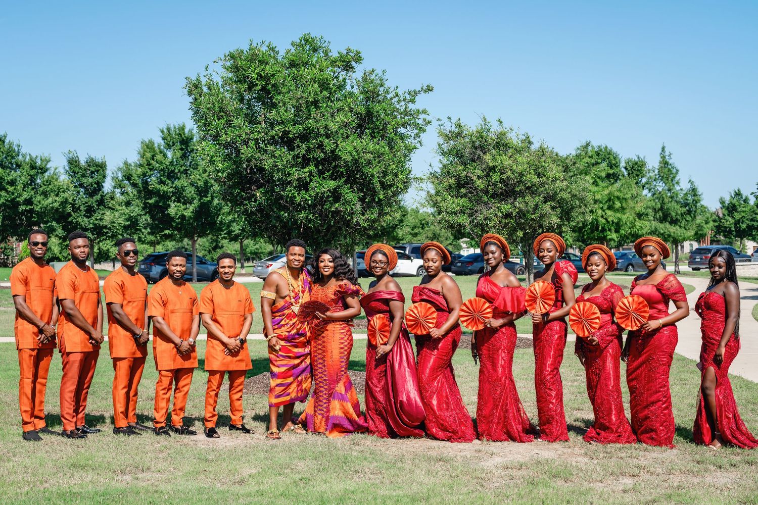 People in vibrant red and orange attire stand in a line formation on green grass under blue skies.