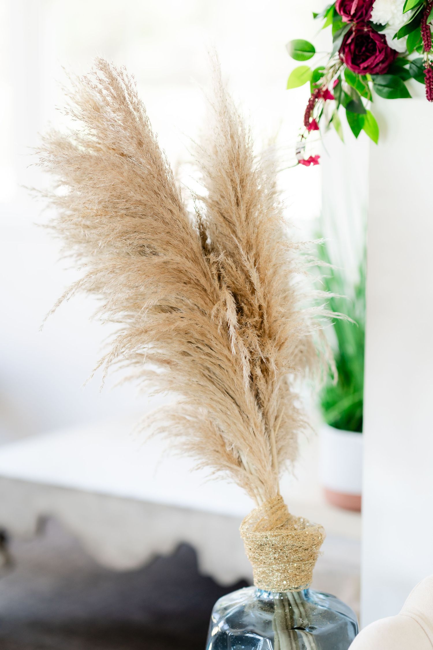 Decorative pampas grass arrangement in a glass vase with burgundy flowers and green foliage.