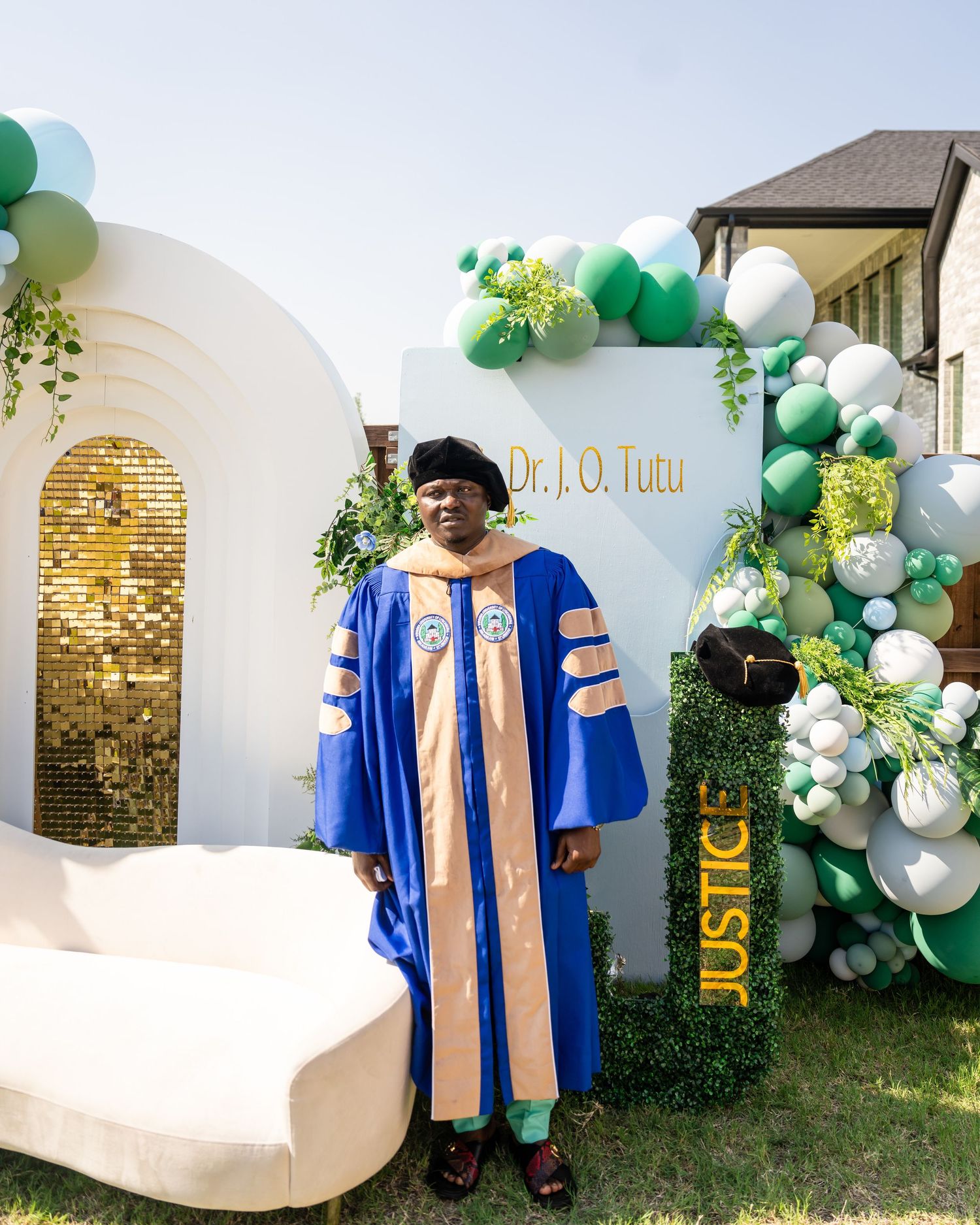 A graduate in royal blue academic regalia poses by a decorative balloon arch with green and white balloons at an outdoor celebration.
