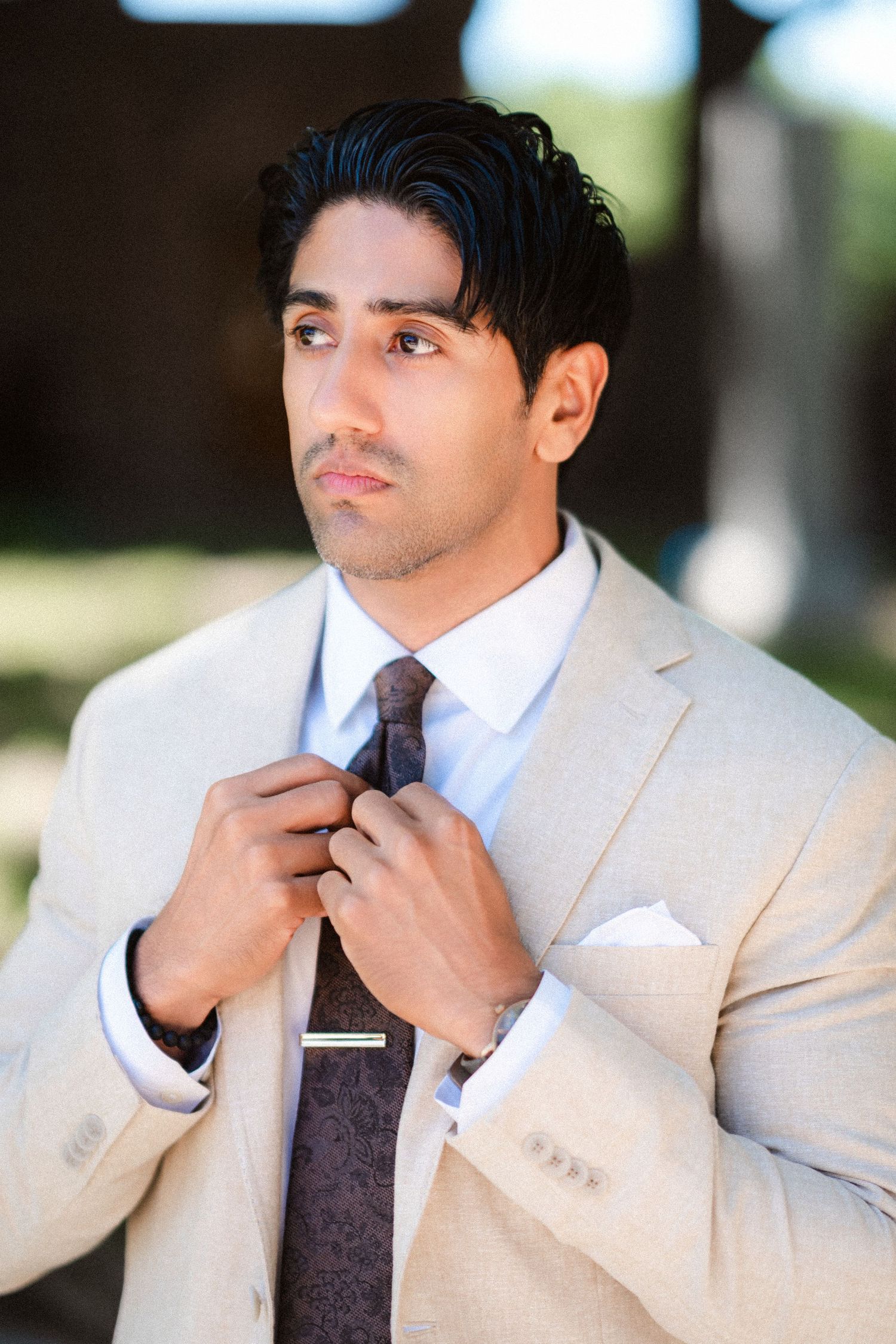 A person adjusts their dark tie while wearing a light colored suit in an outdoor setting.