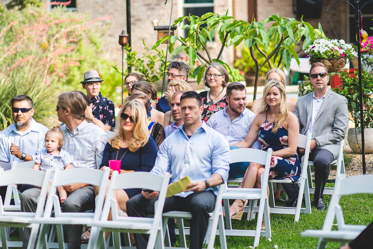 Wedding guests seated in white chairs outdoors during a garden ceremony.