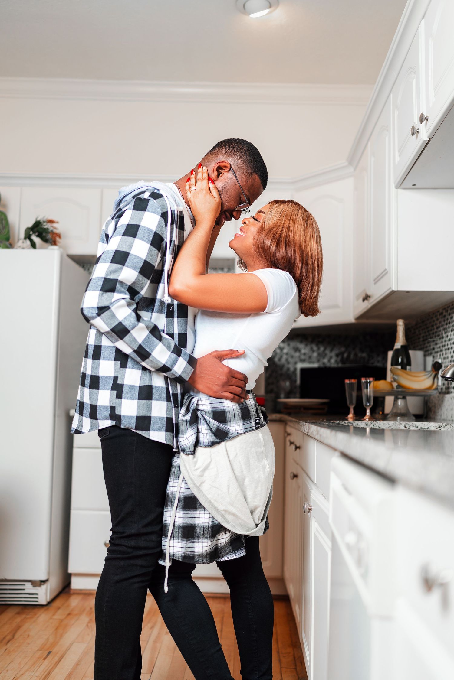 A couple in matching plaid shirts share a tender moment in their modern white kitchen.