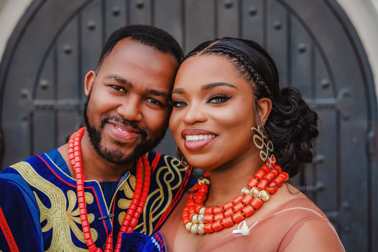 Two people in traditional African attire with colorful beaded necklaces smile together in front of a gray door.