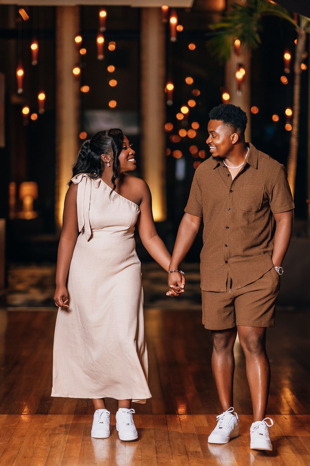 A couple in contrasting brown and white outfits walks together on a warmly lit wooden floor.