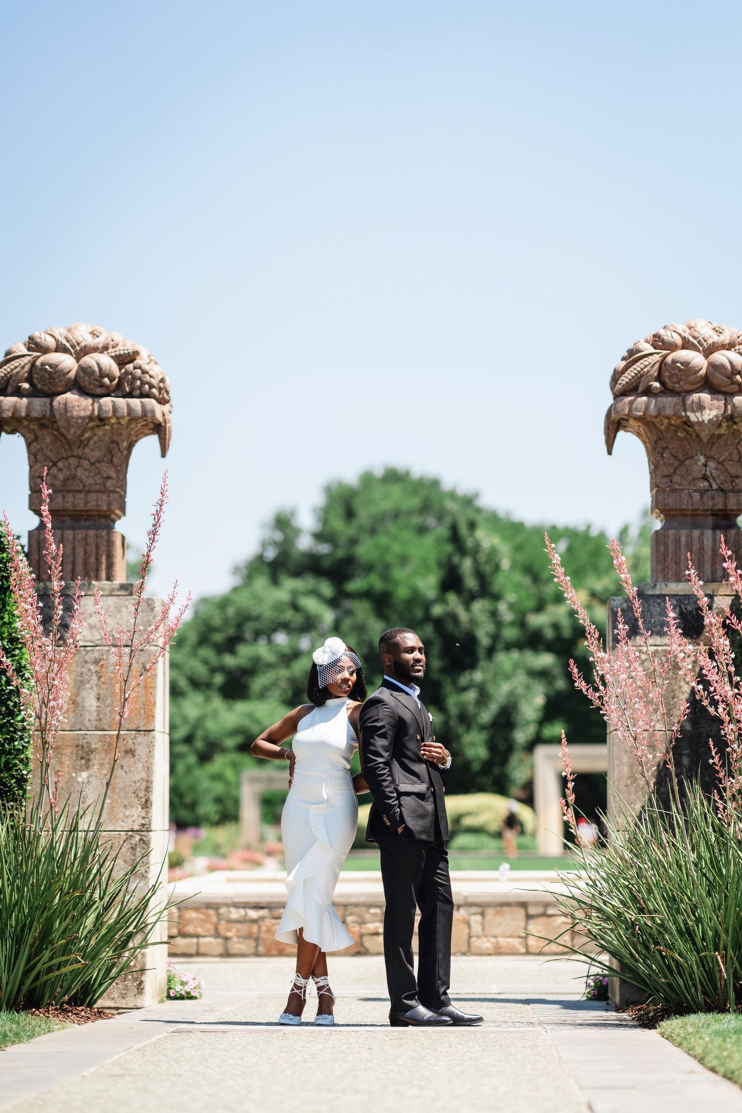 A couple poses between ornate stone pillars in a garden setting wearing elegant wedding attire.