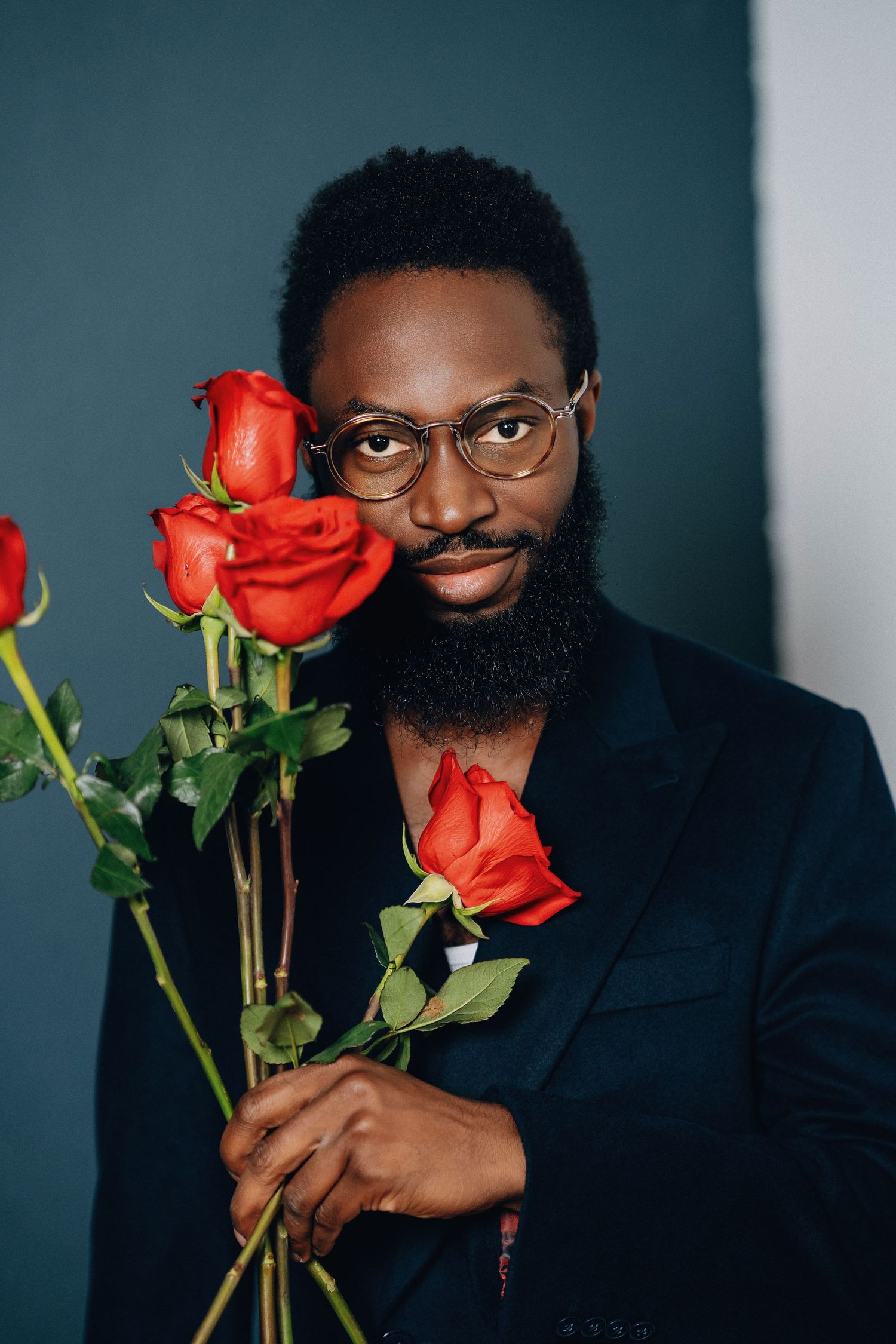 A portrait with red roses against teal background featuring glasses and black blazer.