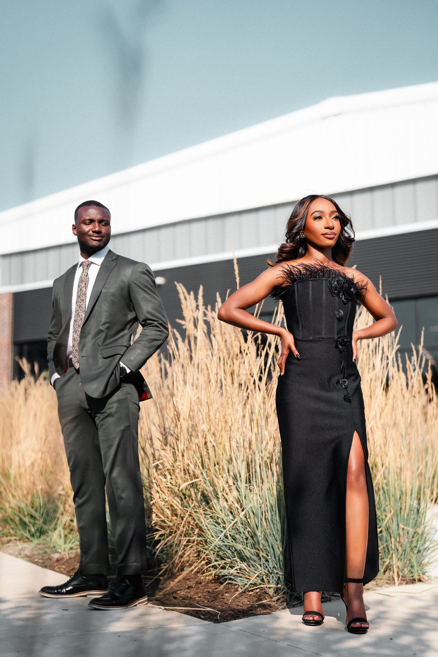 Fashionable couple poses separately against modern building with decorative grasses.