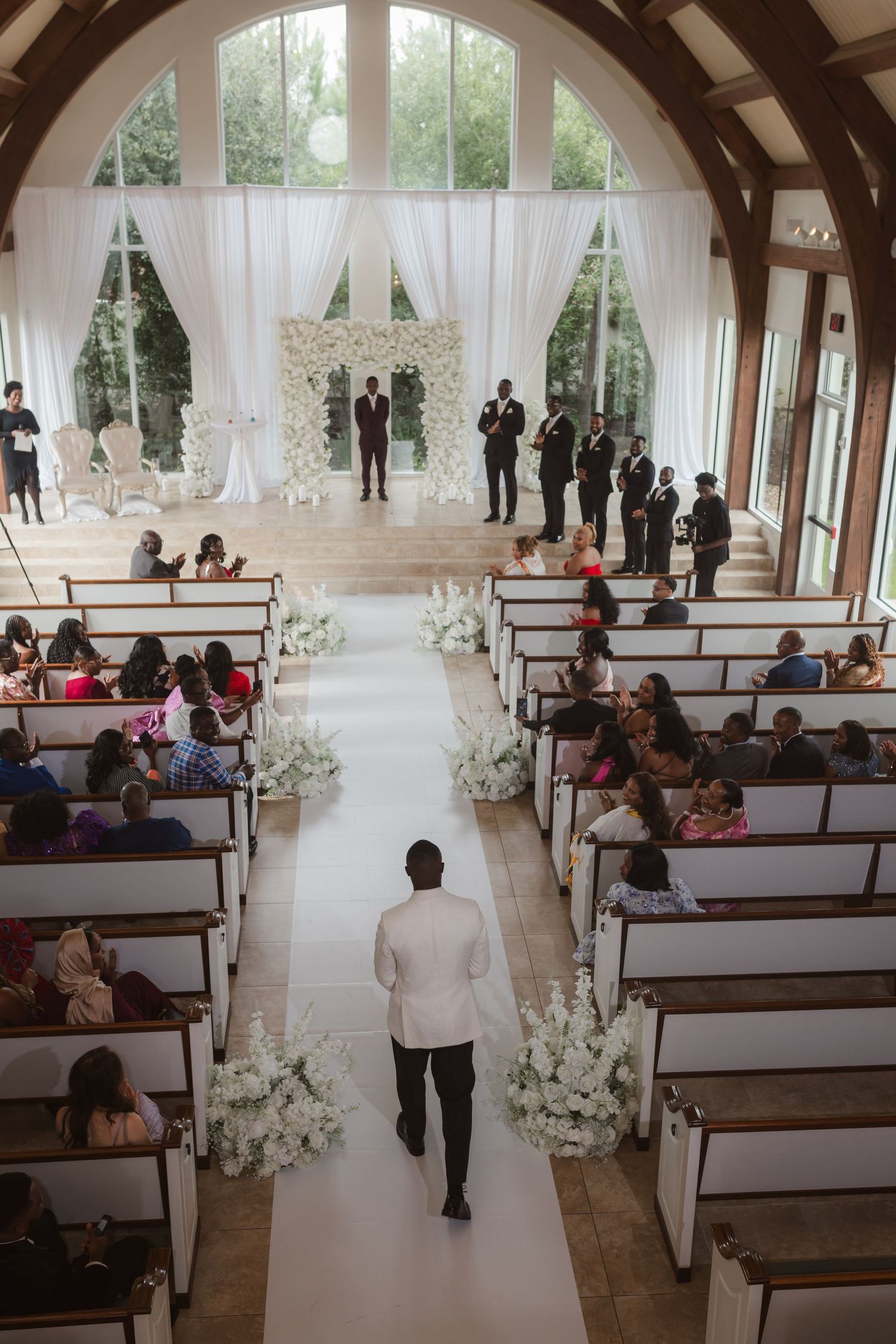 Wedding guests seated in wooden pews decorated with white floral arrangements in a bright indoor venue.