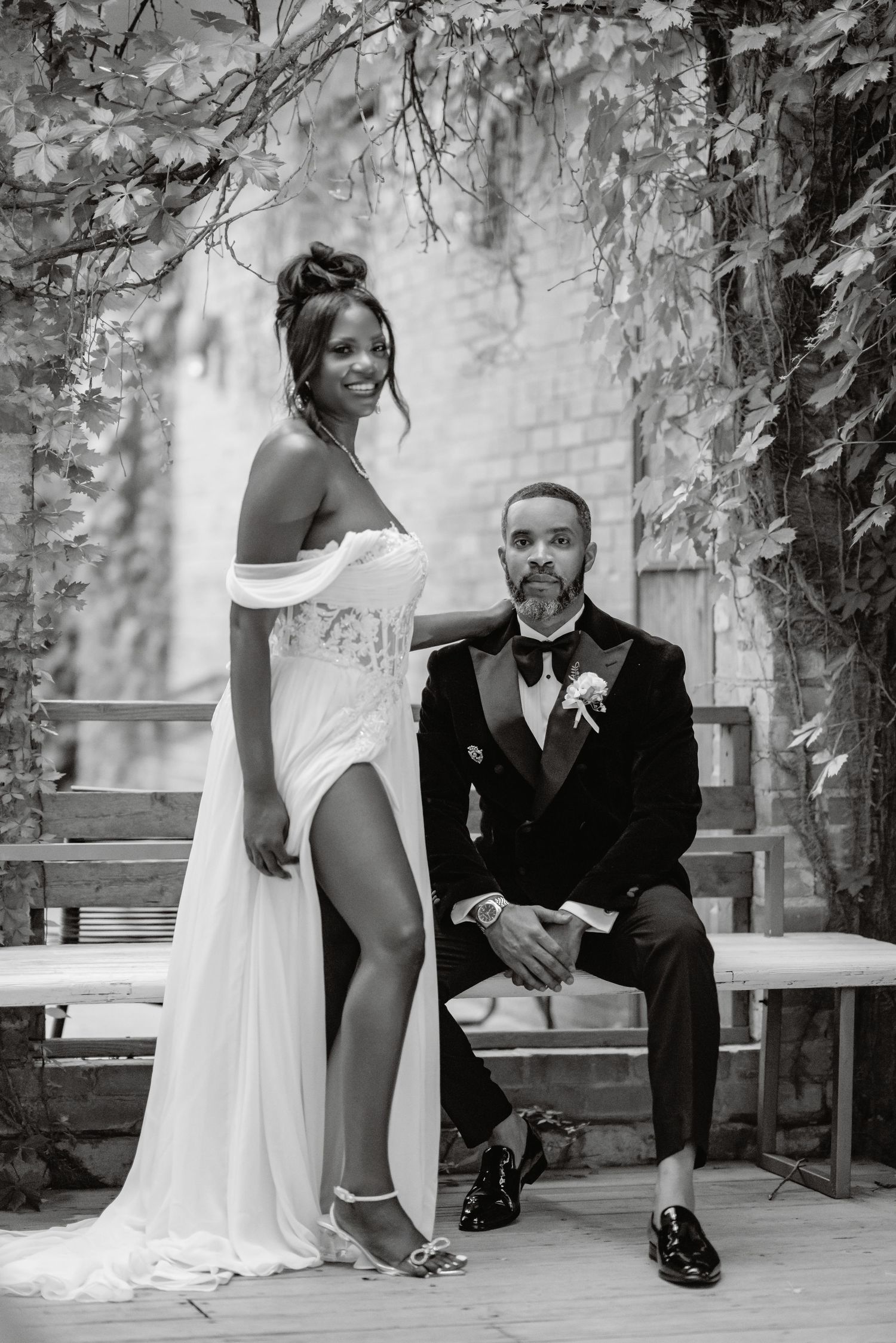 Elegant black and white wedding portrait featuring a bride in a high-slit gown and groom in tuxedo posed in garden setting.