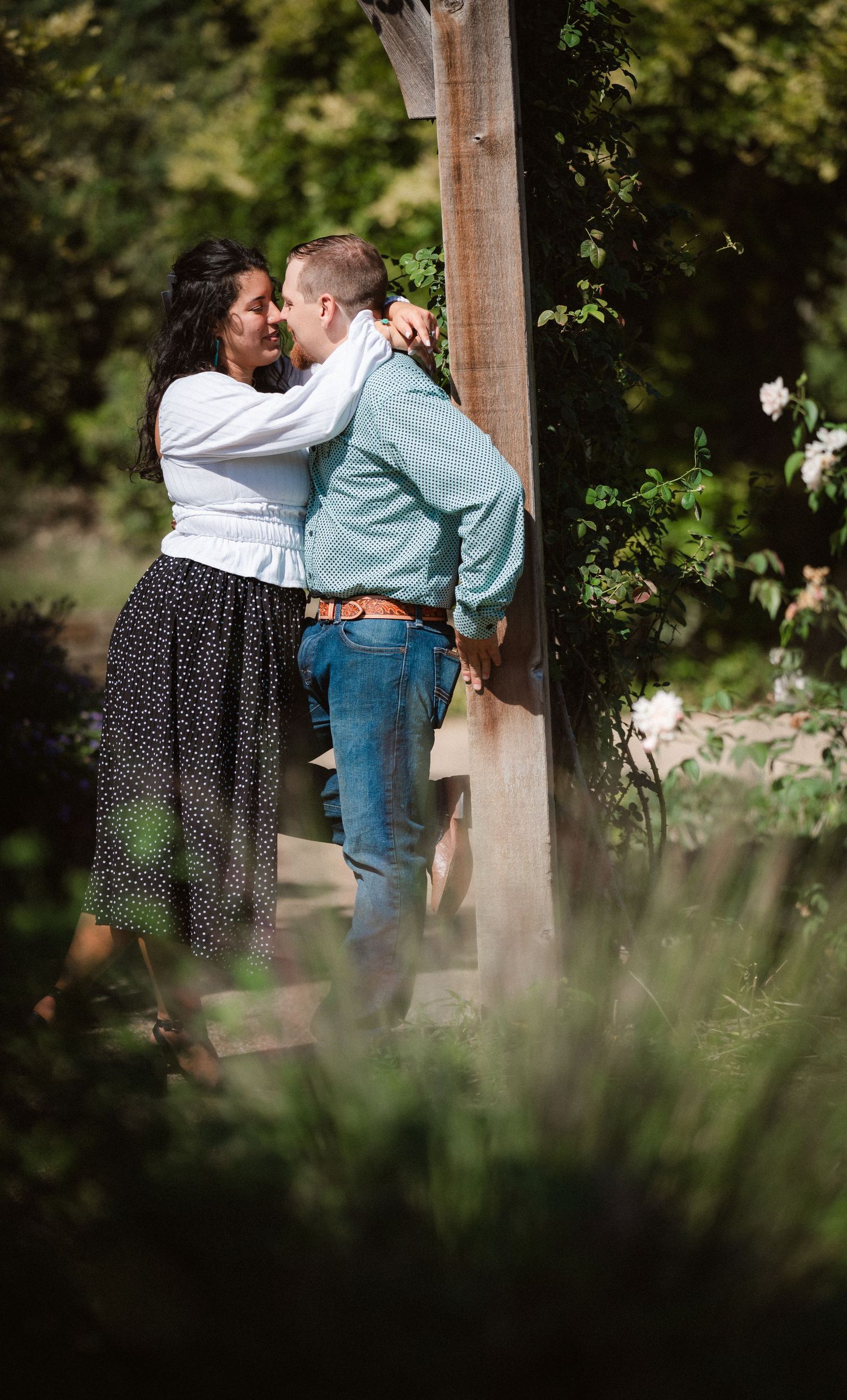 A tender moment captured between a couple in casual attire standing near a wooden post in a lush garden setting.