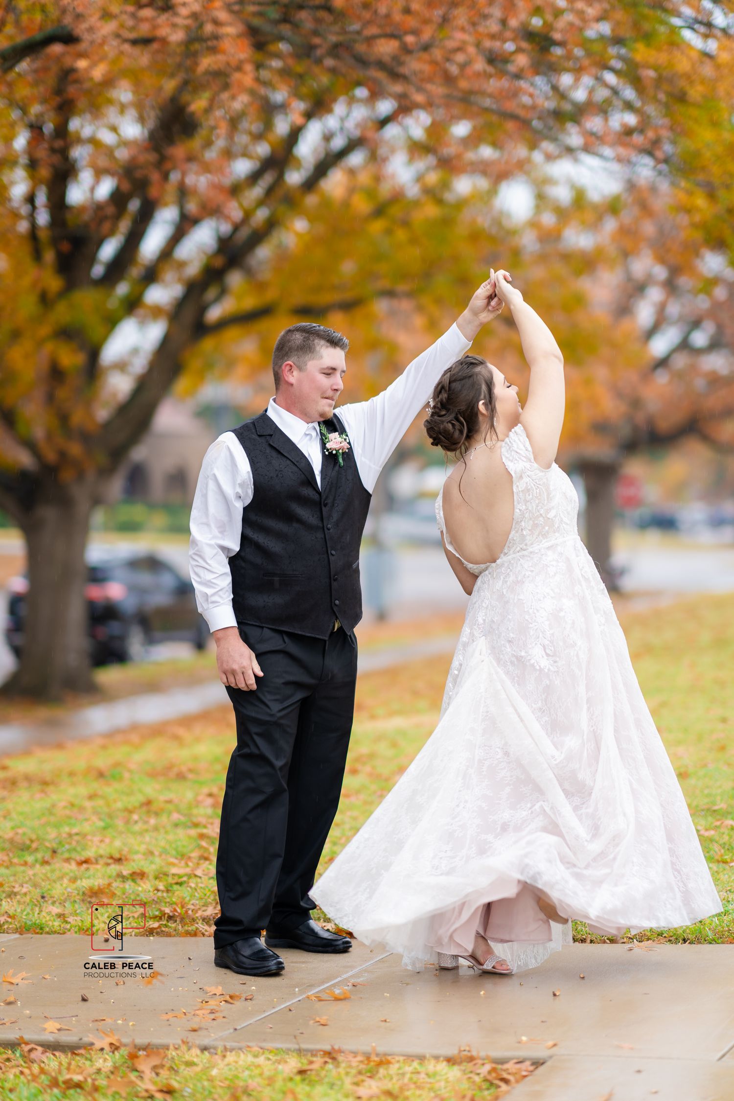 Couple in wedding attire dancing playfully among autumn trees with fallen leaves on the ground.