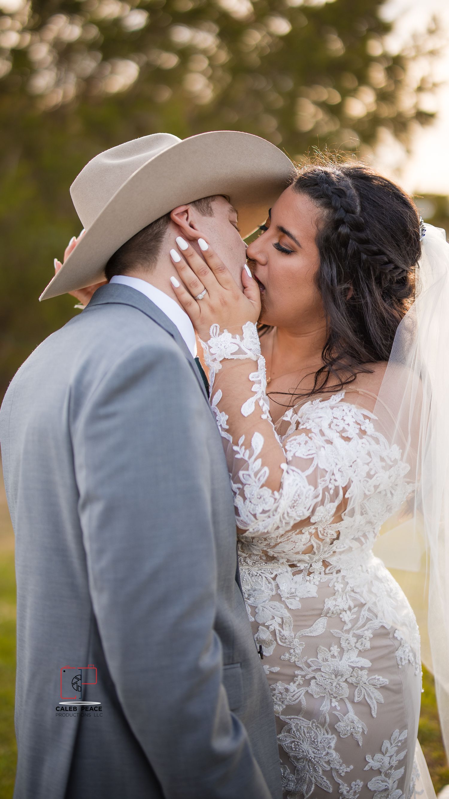 A romantic wedding couple shares a tender kiss outdoors while wearing a grey suit and lace gown with veil.