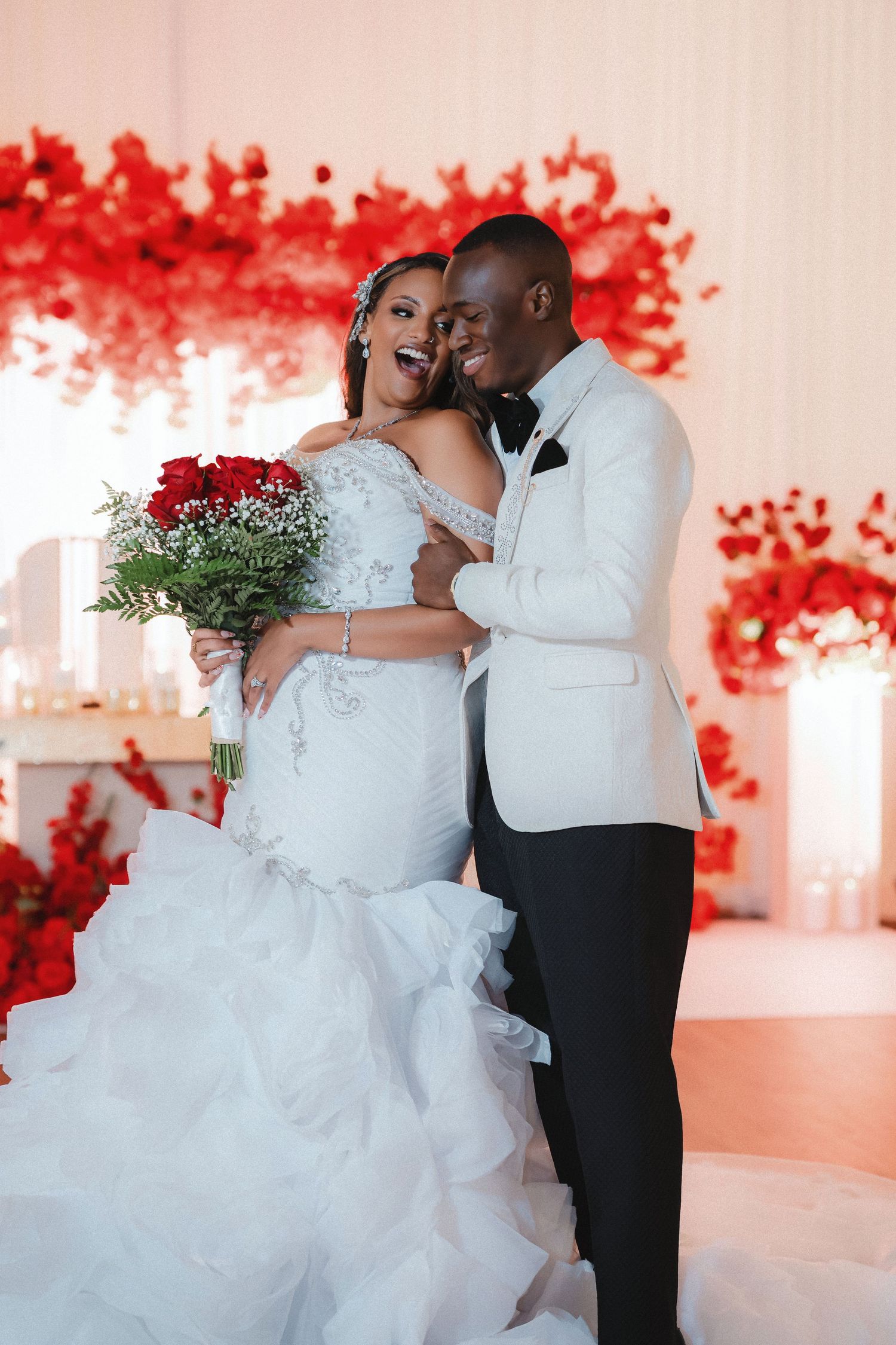 A couple shares a romantic dance moment in a wedding reception venue with red floral decor and white walls.