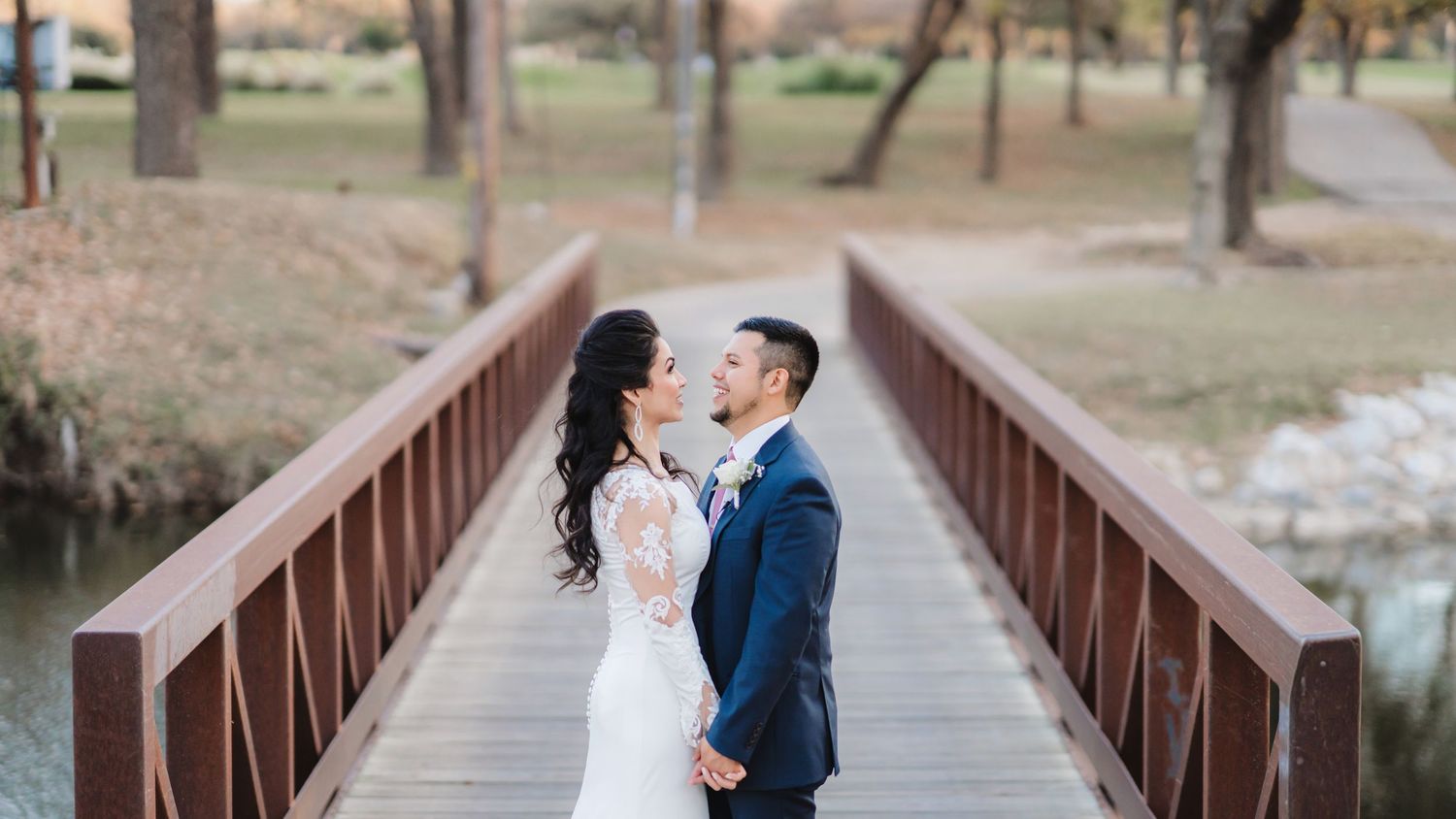 A wedding couple walks hand in hand across a wooden bridge surrounded by nature.