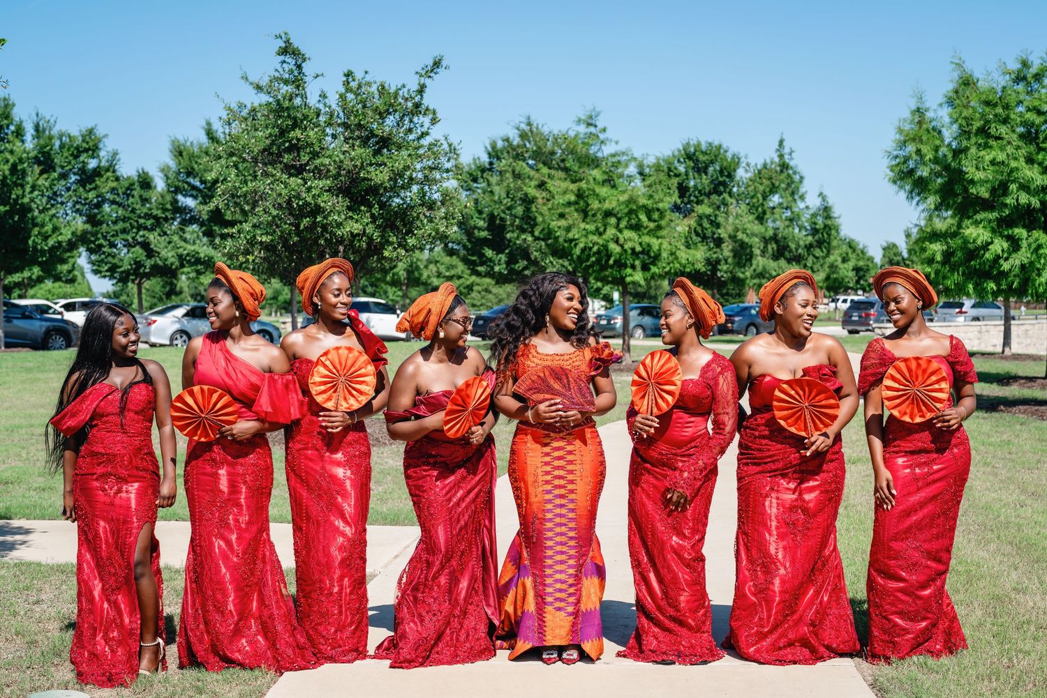 Wedding party wearing matching red dresses poses in a line outdoors on a sunny day.