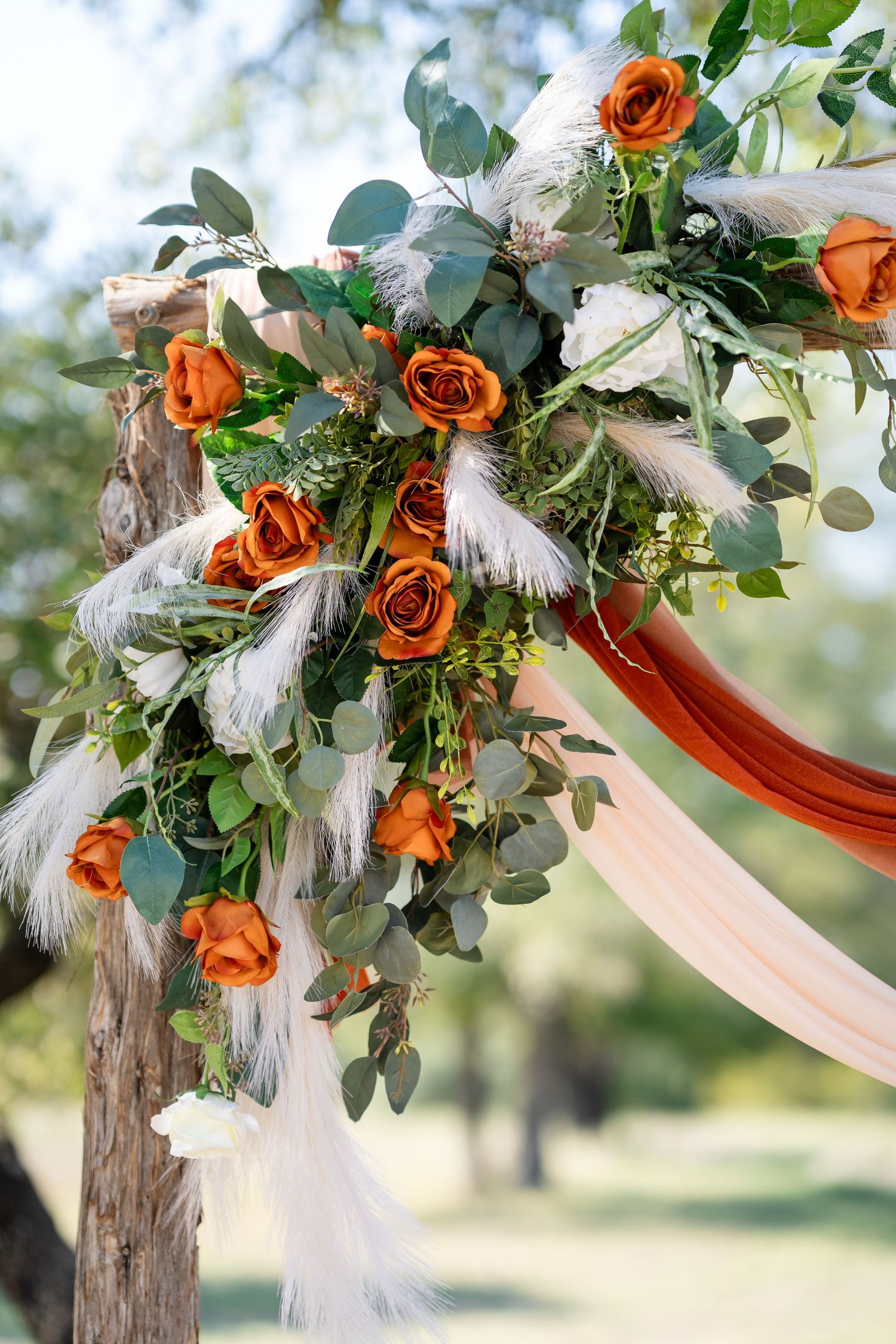Wedding arch decoration featuring orange roses, eucalyptus leaves, and white feathers against a blurred outdoor background.