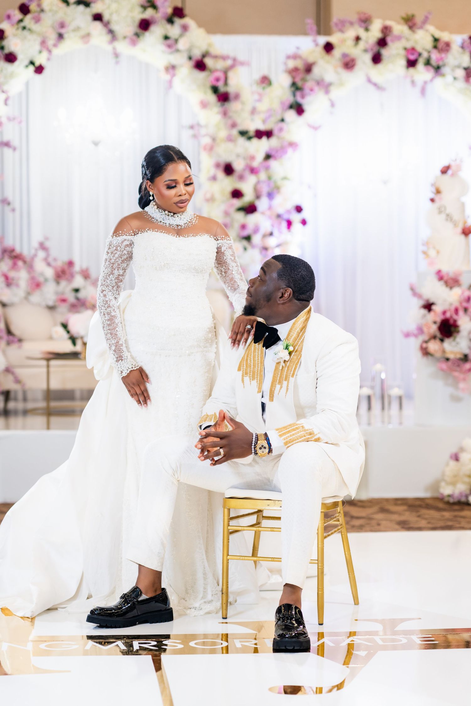 Elegant wedding couple shares intimate moment while seated on gold chairs against floral wall.