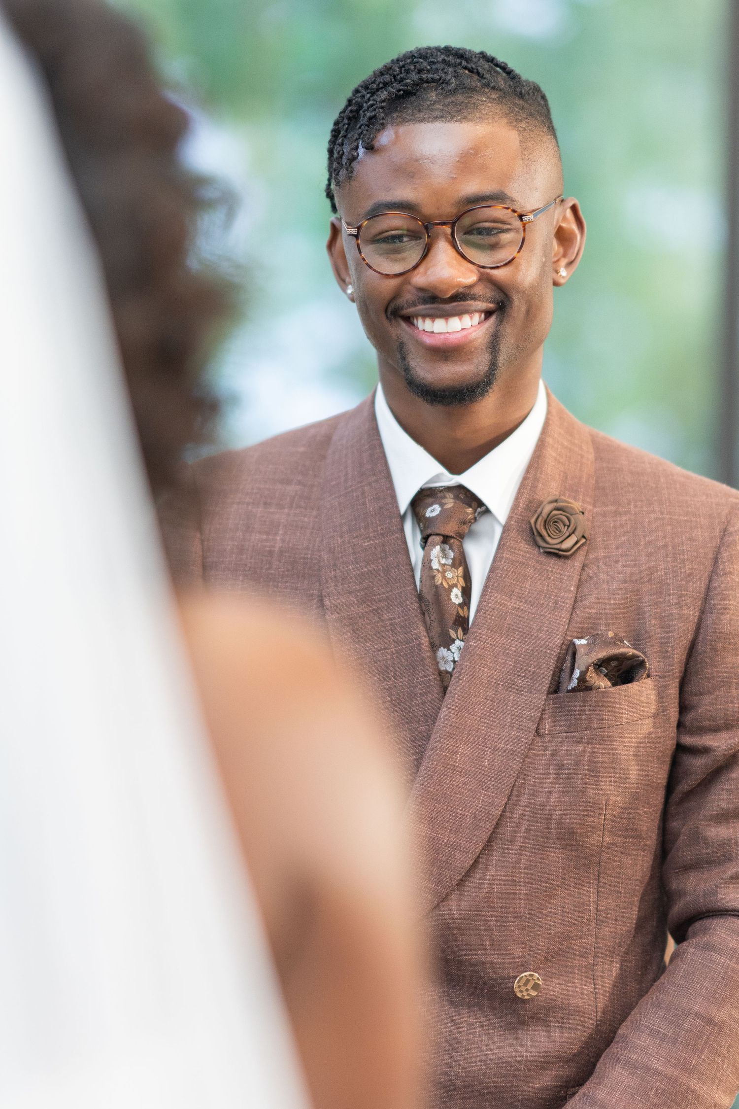 Groom wearing a stylish brown suit and glasses smiles during wedding ceremony.