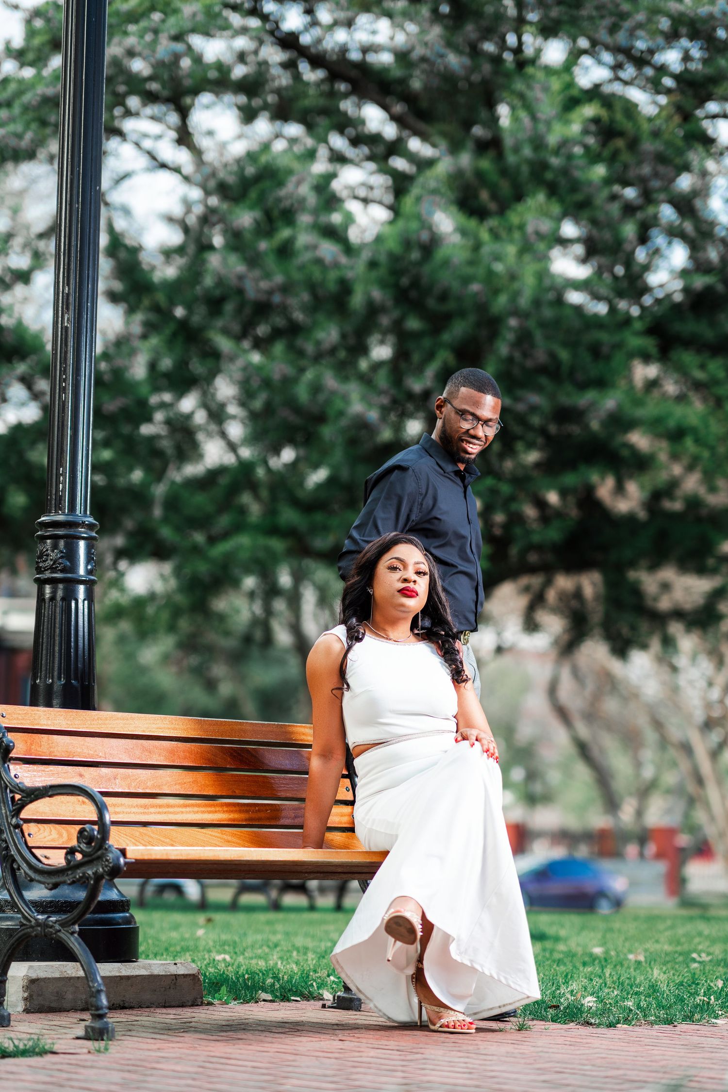 A romantic couple poses on a park bench near a vintage lamppost surrounded by lush greenery.