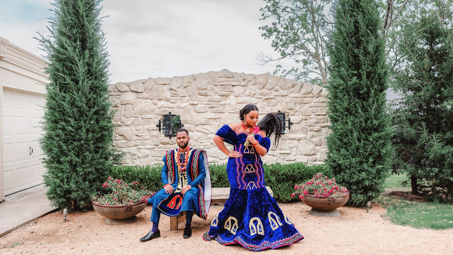 A couple in traditional blue and red formal attire pose playfully in front of stone archways.