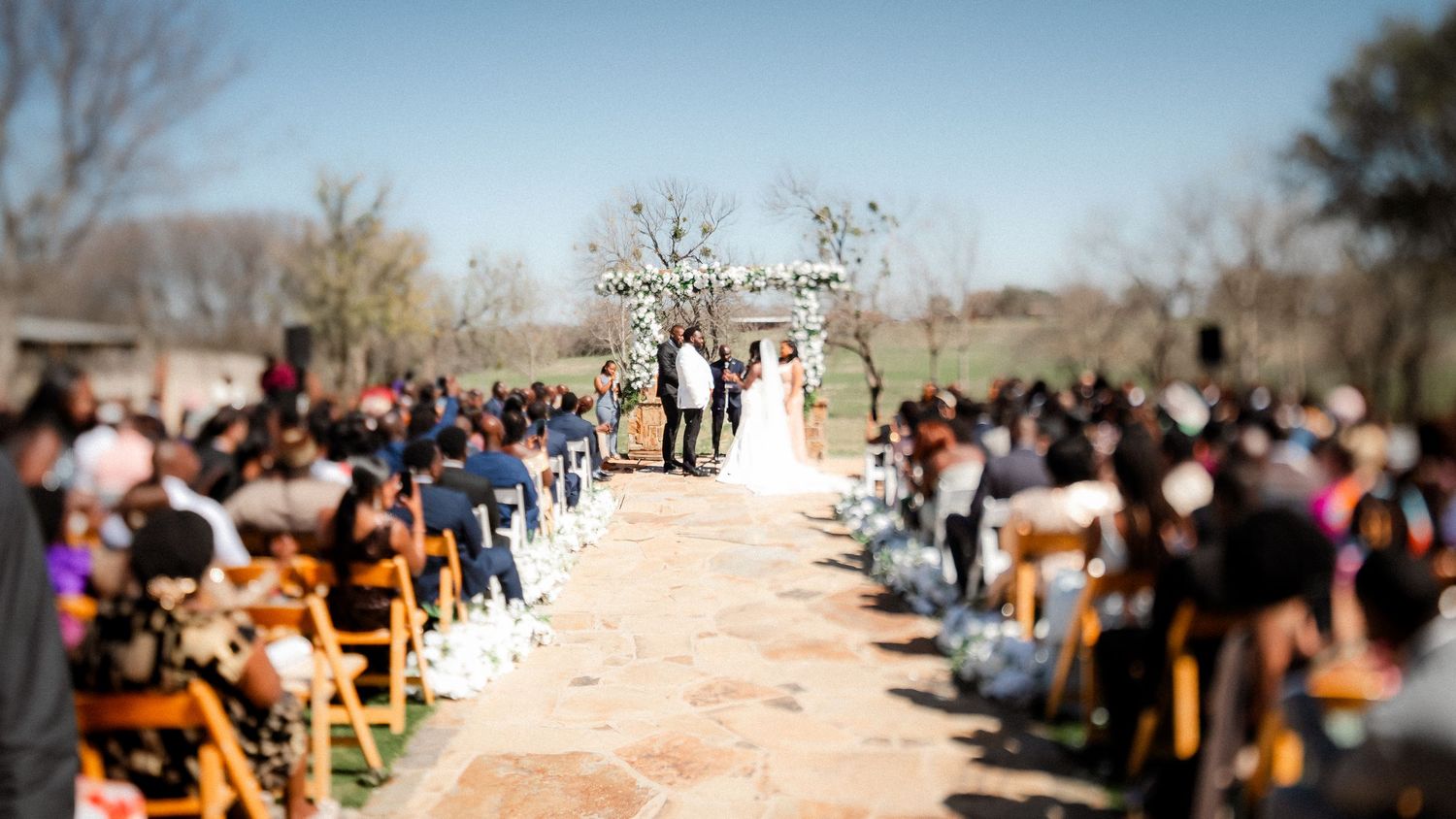 An outdoor wedding ceremony with guests seated on wooden chairs along a stone pathway under a bright blue sky.