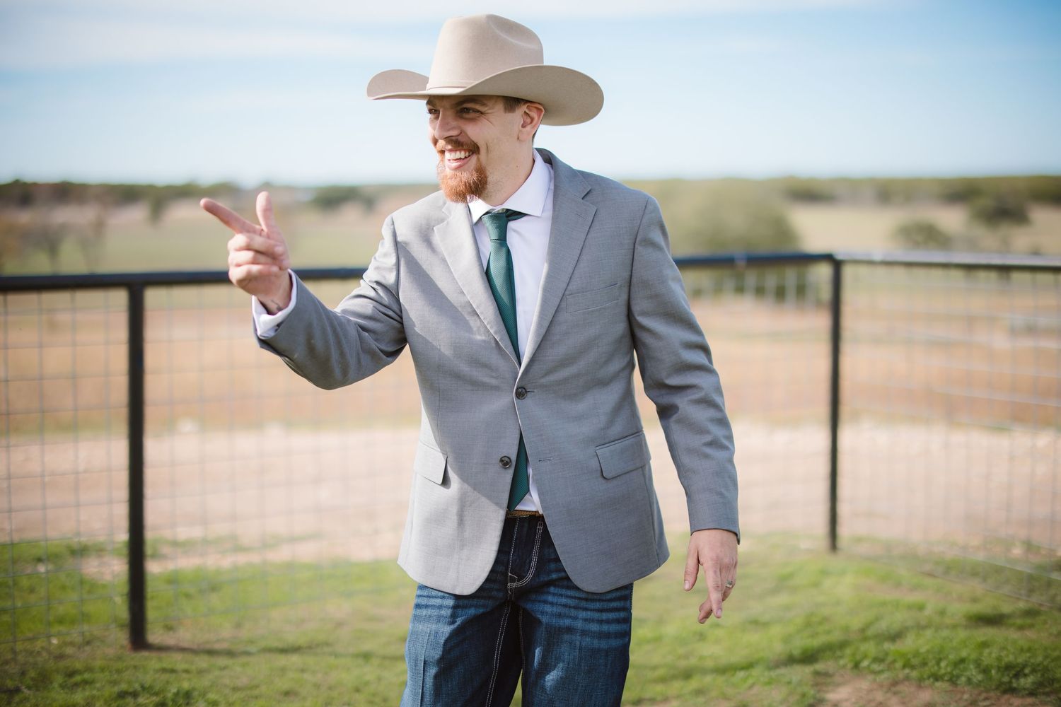 A person in a gray suit jacket and cowboy hat stands in a sunny field near a wooden fence.