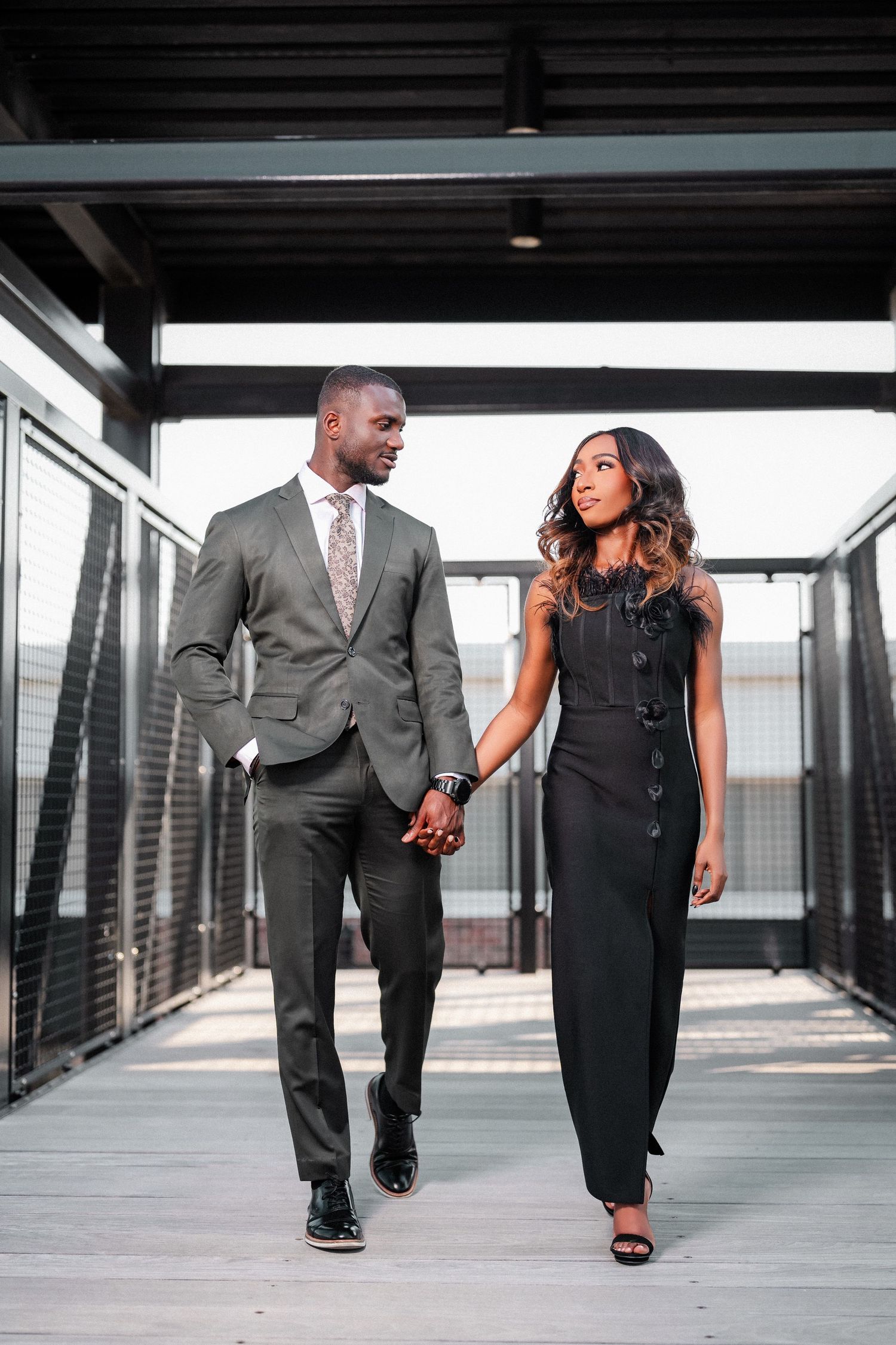 A stylish couple walks down a modern hallway in coordinated black and grey formal attire.