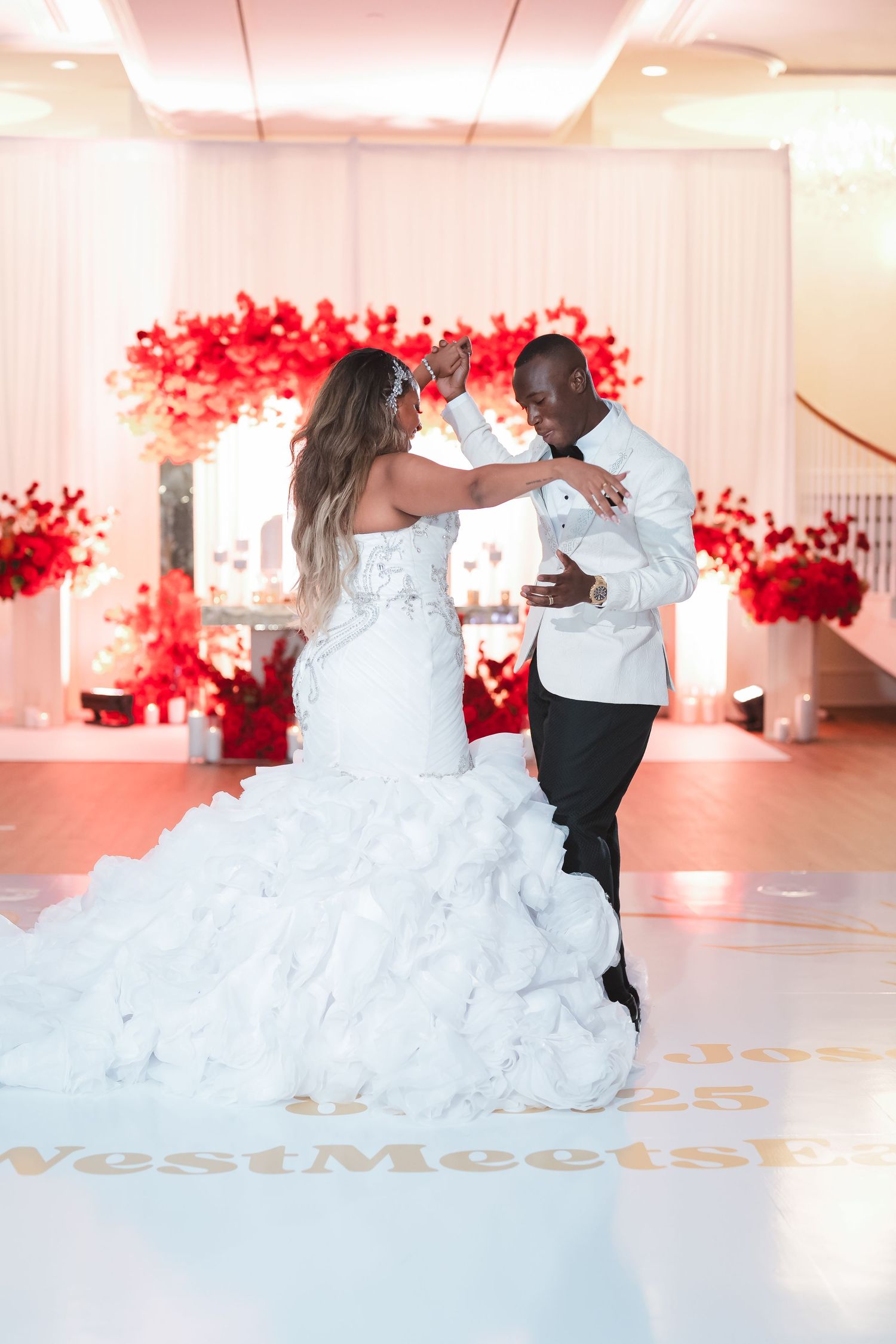 A couple shares their first dance in a reception hall decorated with red floral accents.