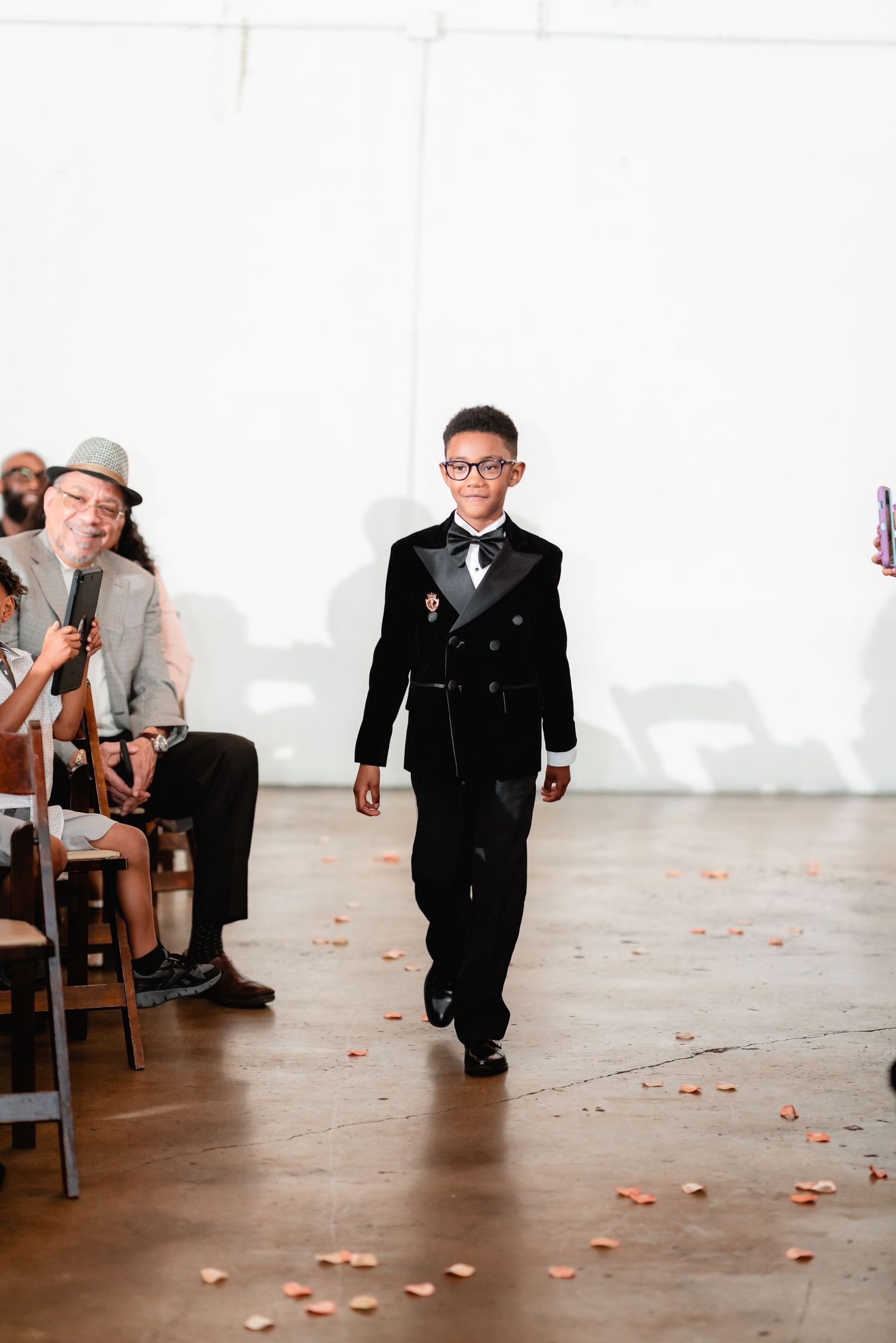 Ring bearer walks down aisle scattered with rose petals during indoor wedding ceremony.