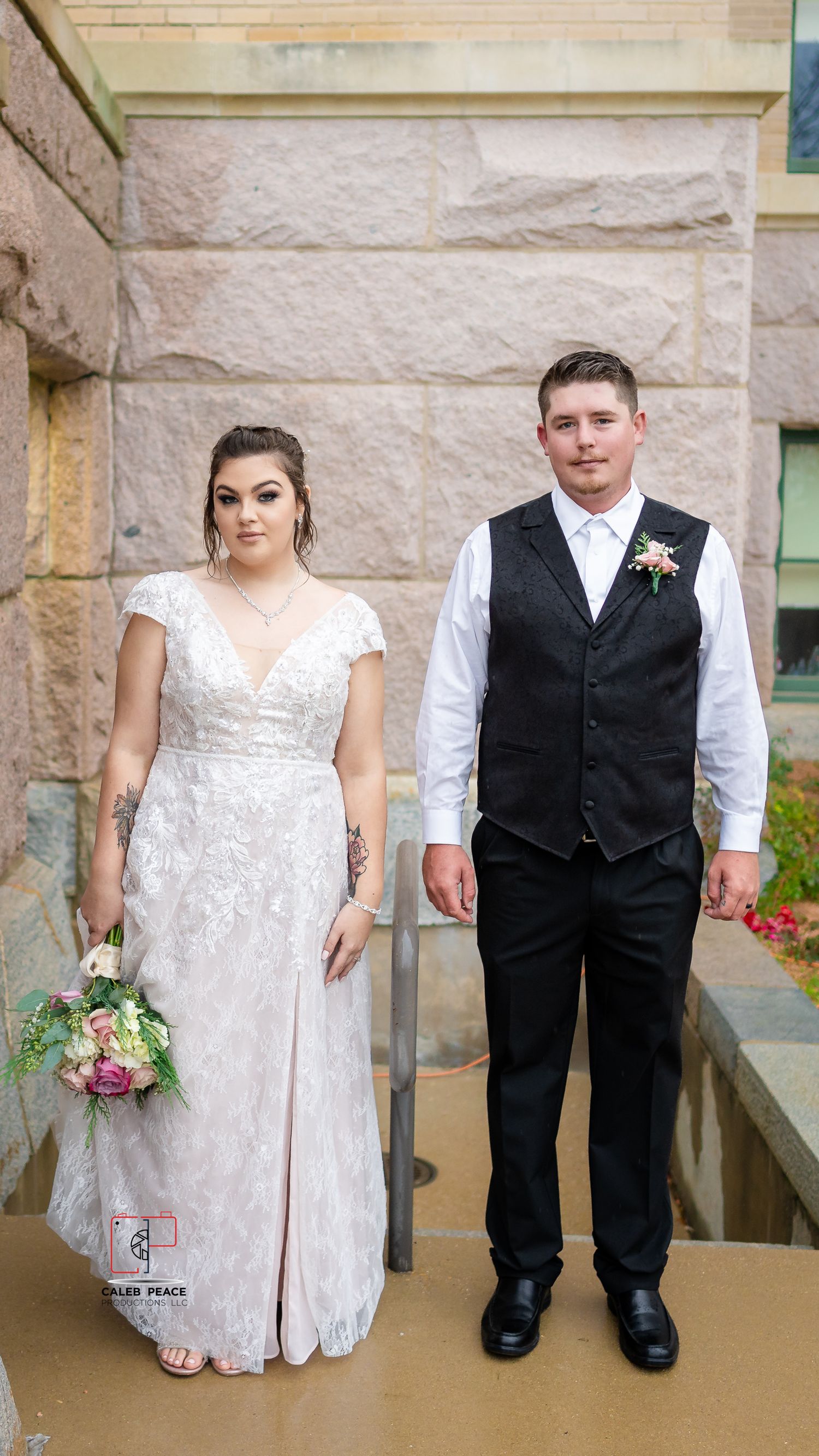 A bride in a lace wedding dress and groom in a black vest stand together on stone steps during their wedding ceremony.