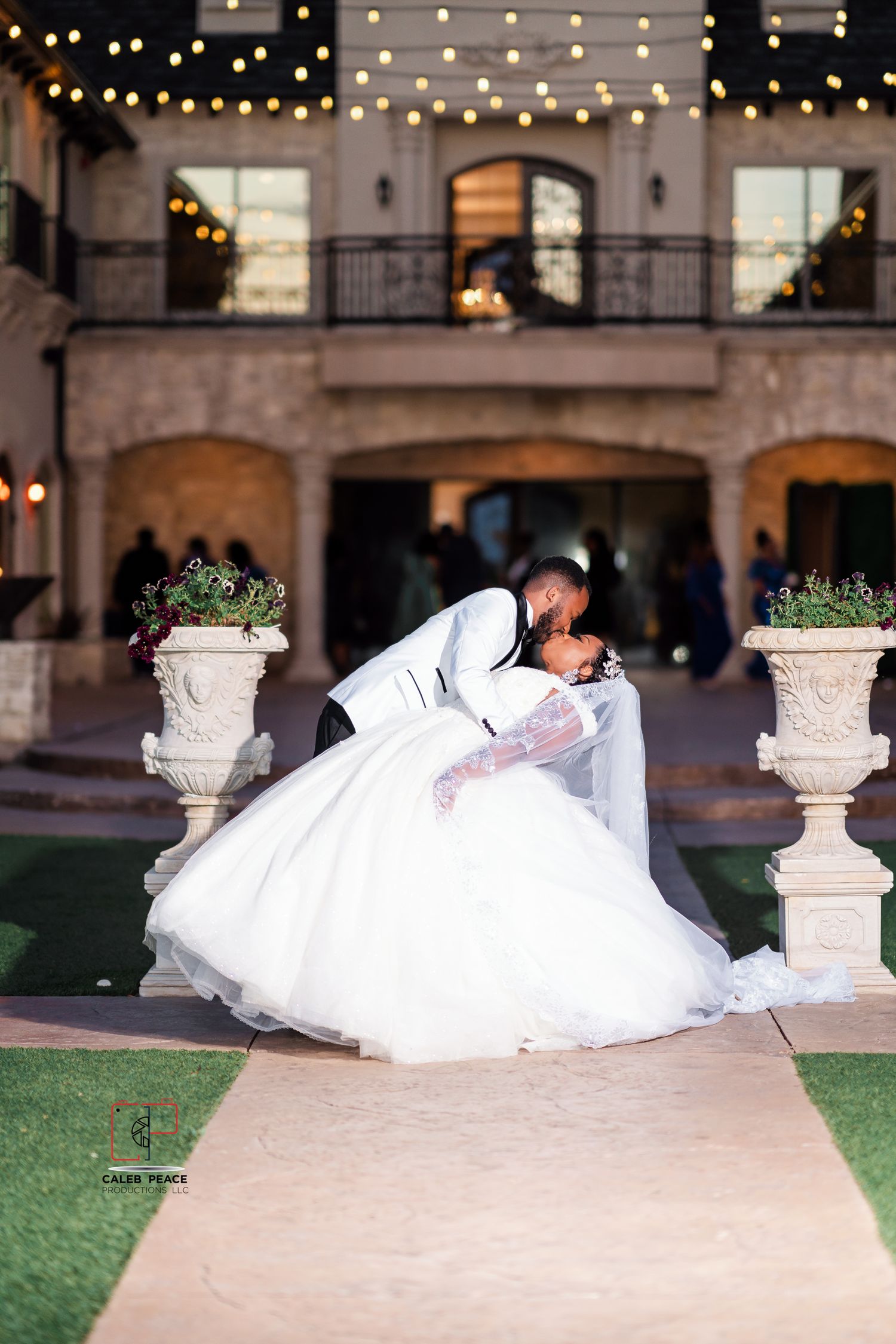 A romantic wedding couple lies on grass near stone pillars with string lights glowing in the background.