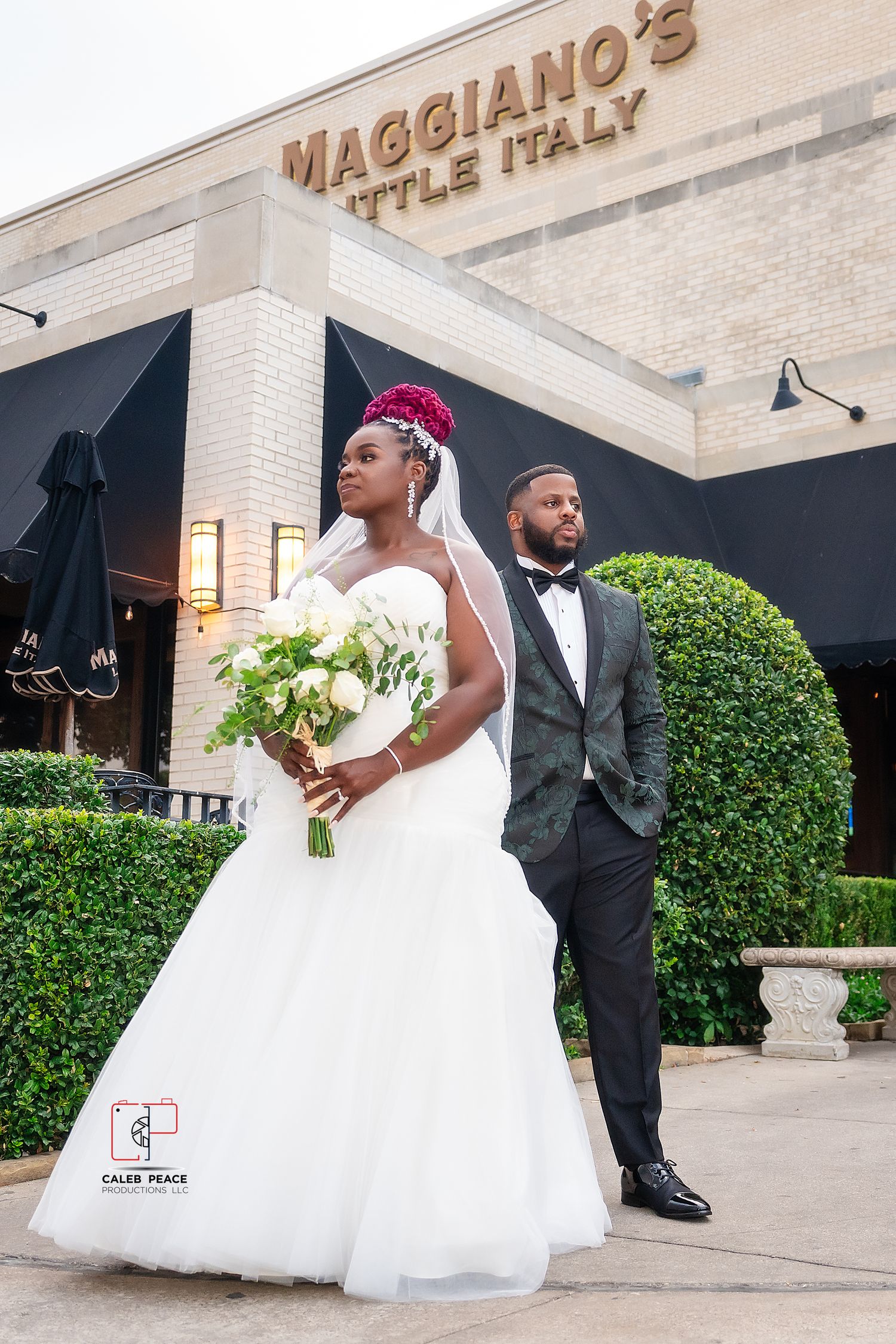 A couple in wedding attire poses outside Maggiano's restaurant with black umbrellas and white flowers.