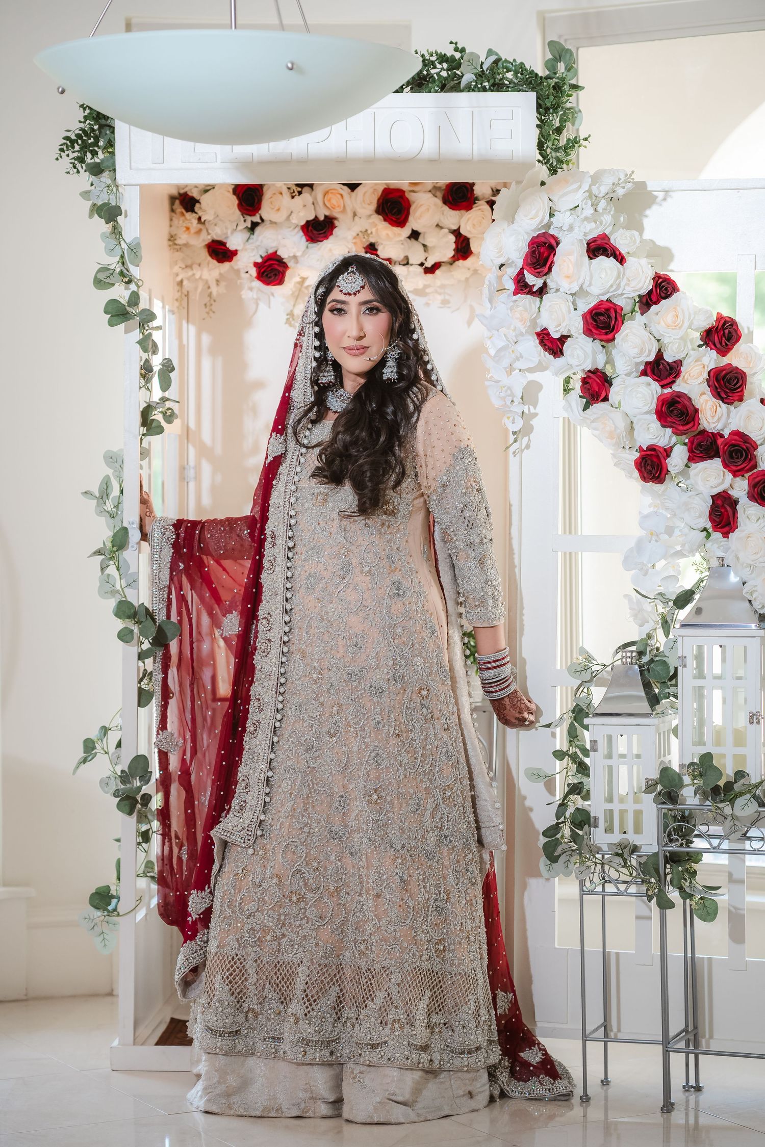 Elegant bridal attire featuring a beaded ivory lehenga with red rose decor and greenery in a light-filled room.