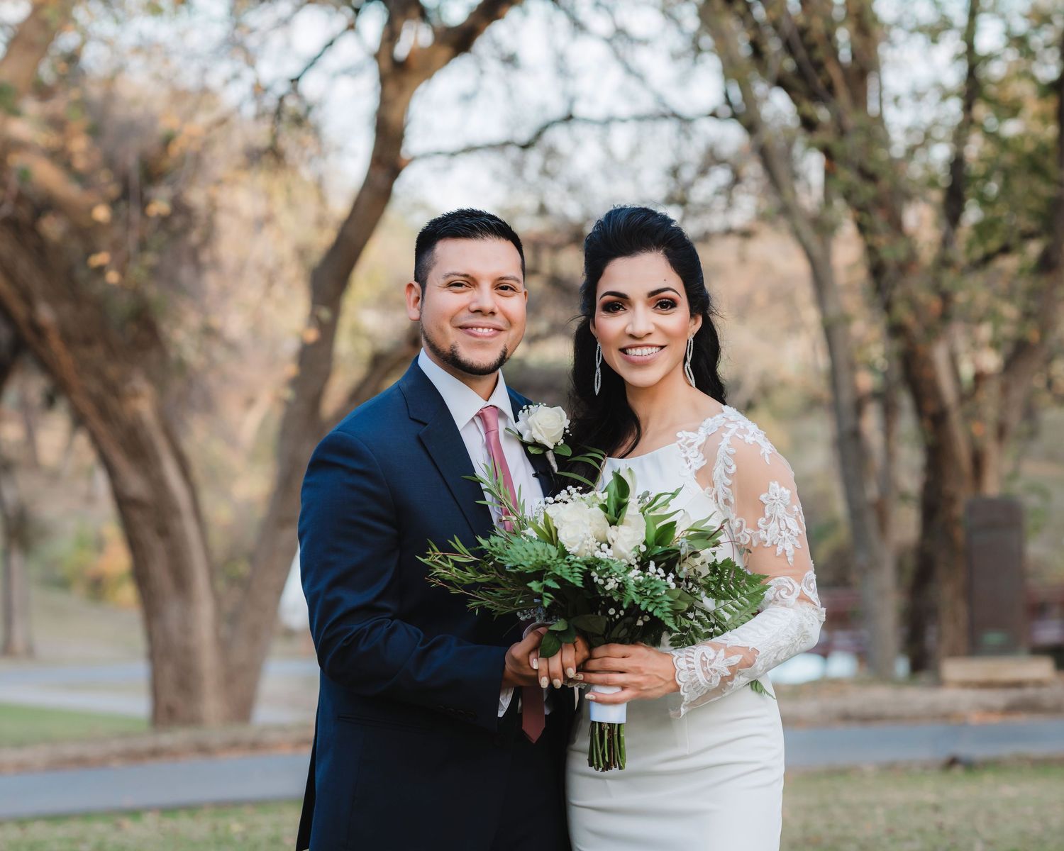 A couple in wedding attire embrace outdoors among autumn trees with the bride holding a green bouquet.