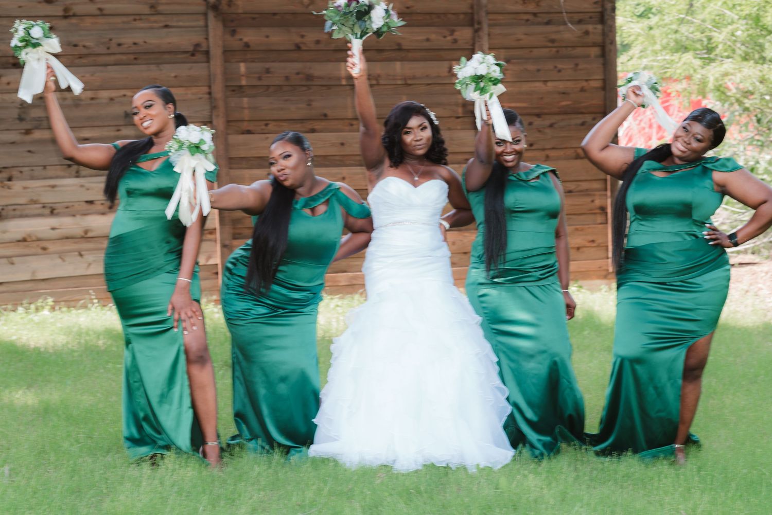 Wedding party poses in coordinated emerald green dresses on grassy lawn.