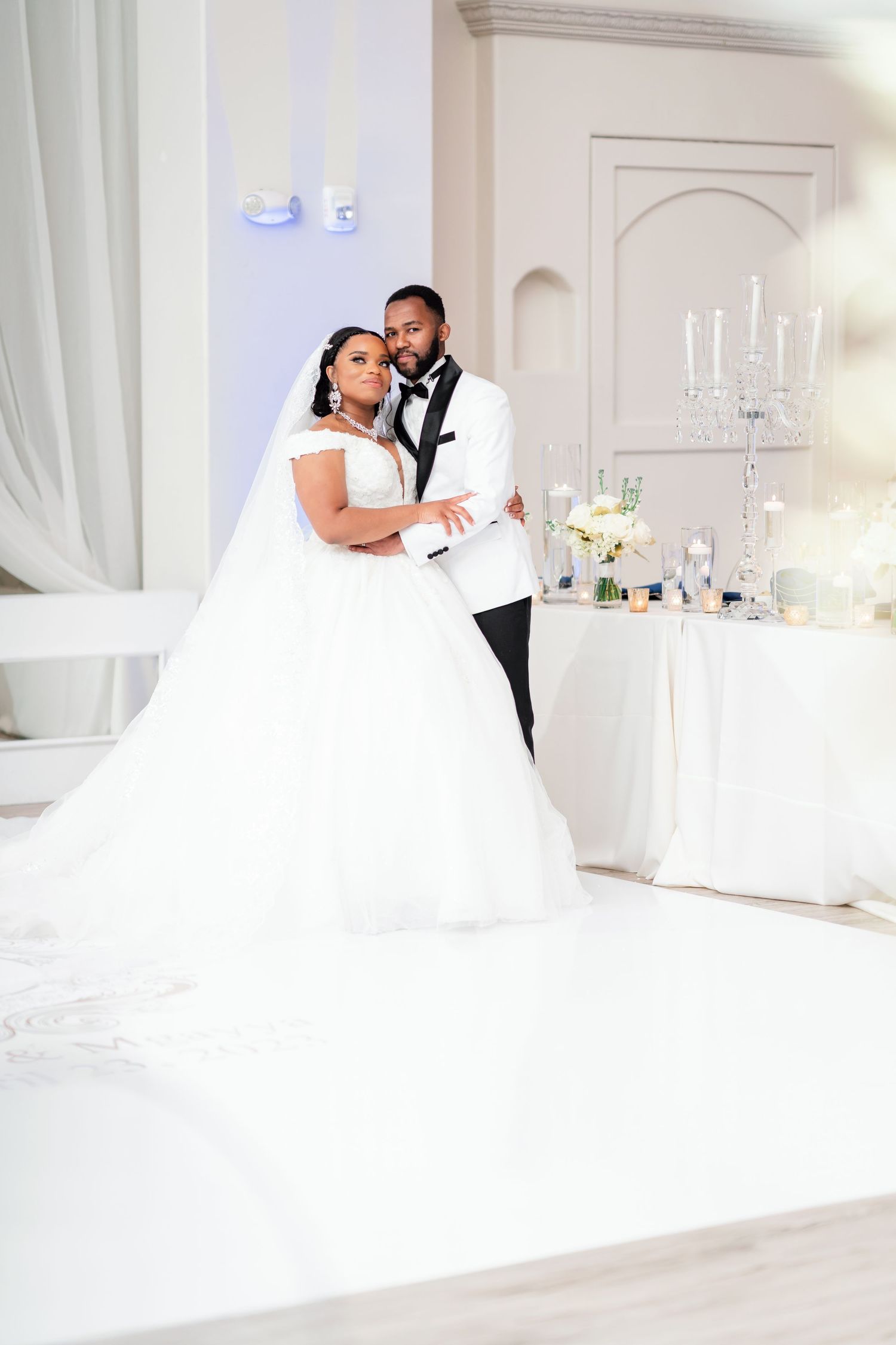 Elegant wedding couple embraces in an all-white luxury reception venue with candelabras and flowing curtains.