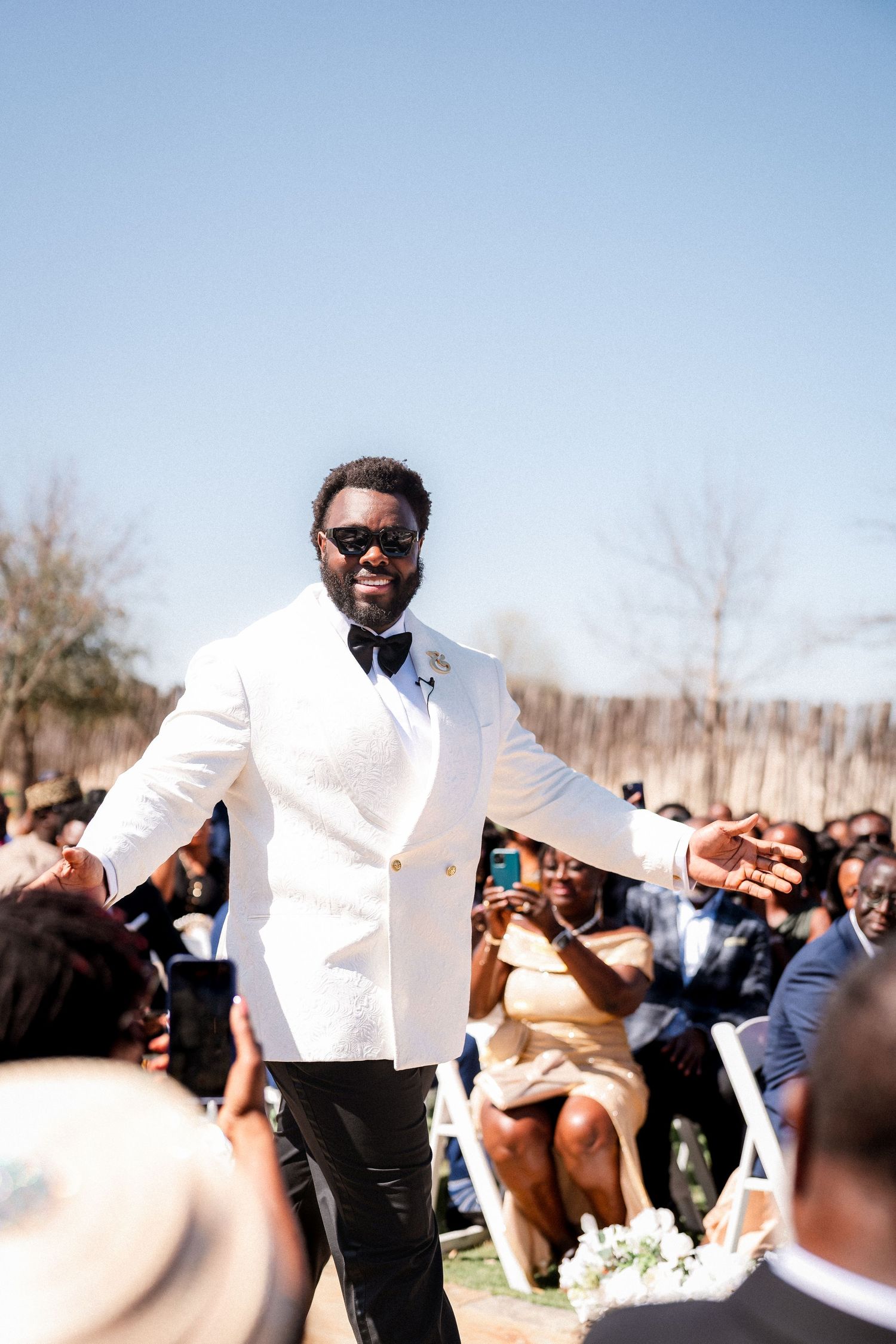 A groom in a white tuxedo celebrates with wedding guests during an outdoor reception.