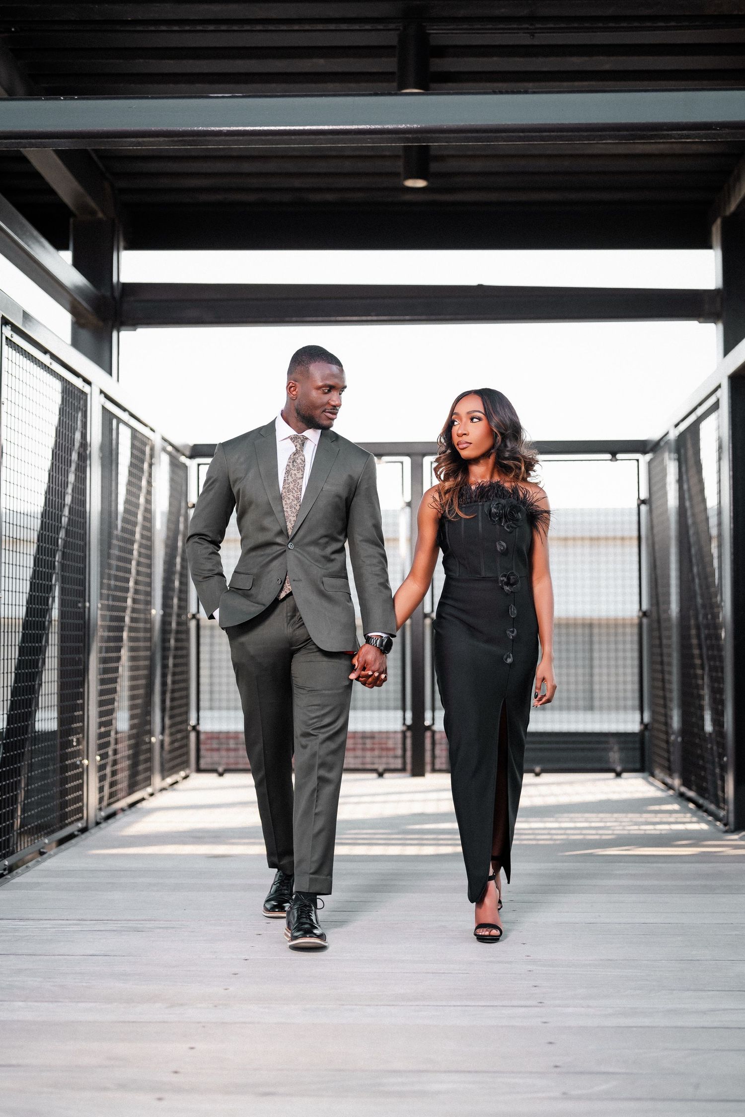 Stylish couple in formal attire walking together through a modern hallway holding hands.