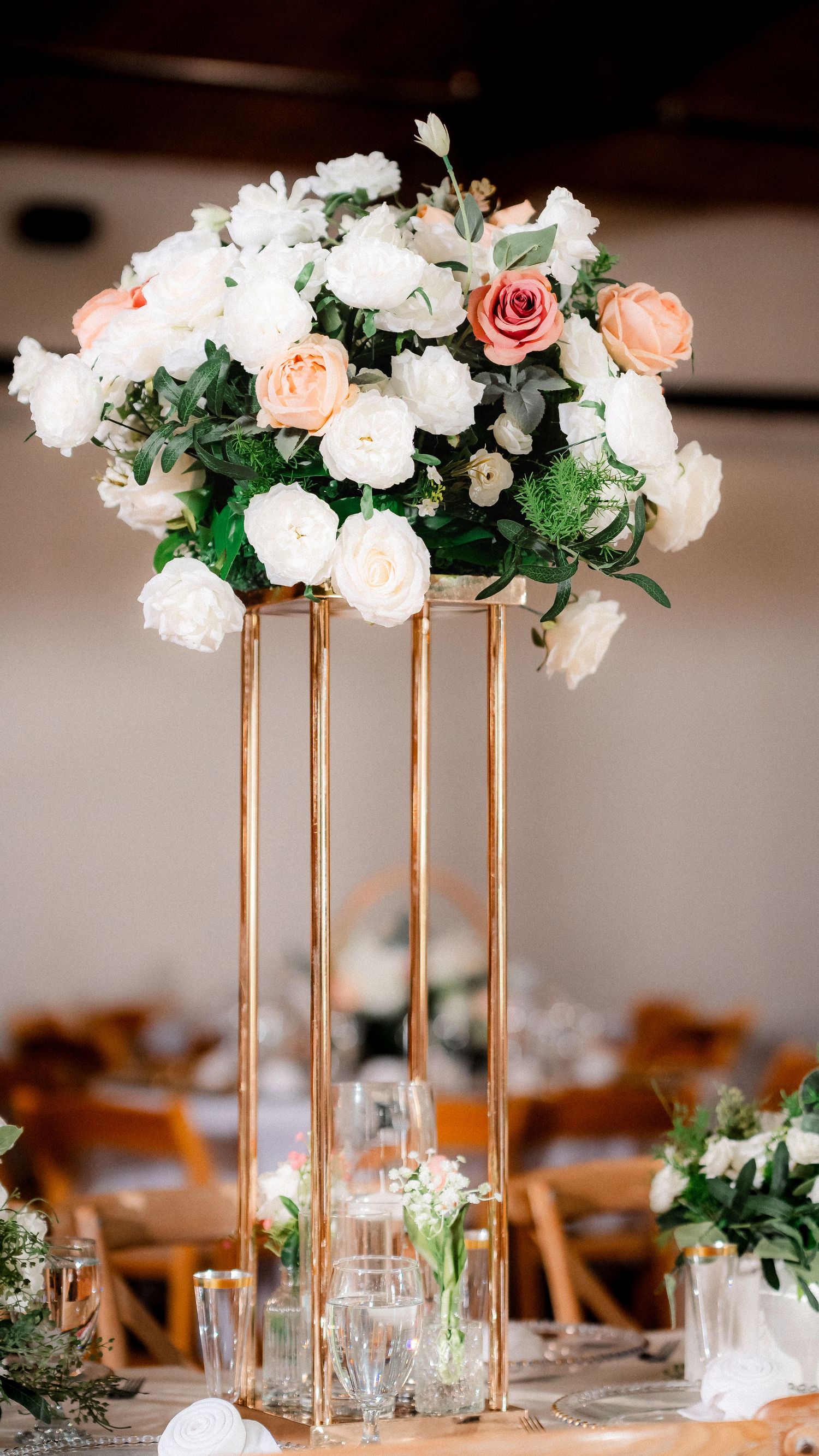 Elegant wedding centerpiece with white and peach roses arranged on tall gold stands at reception tables.