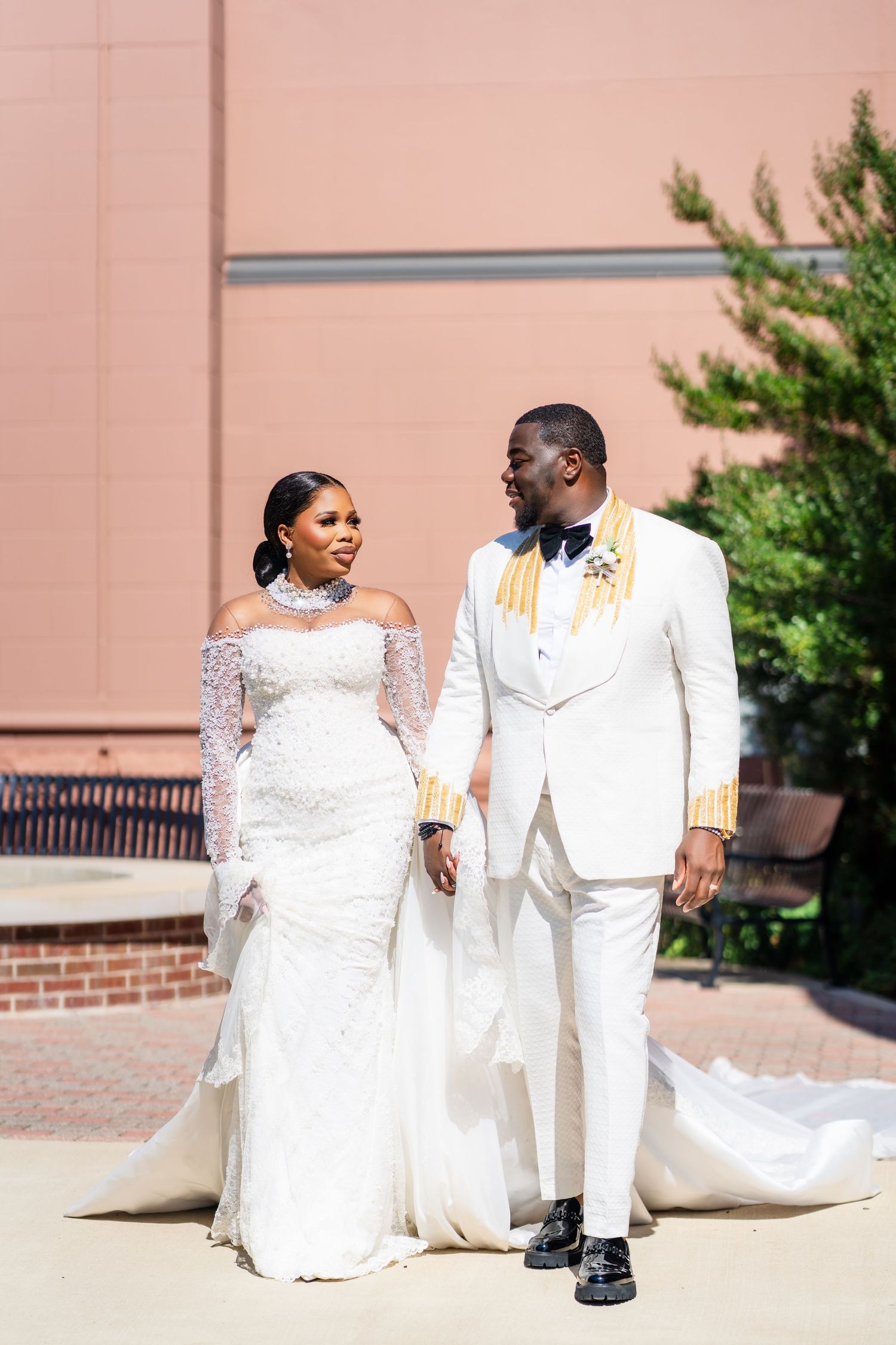 A couple in elegant white wedding attire poses together outdoors against a pink wall.