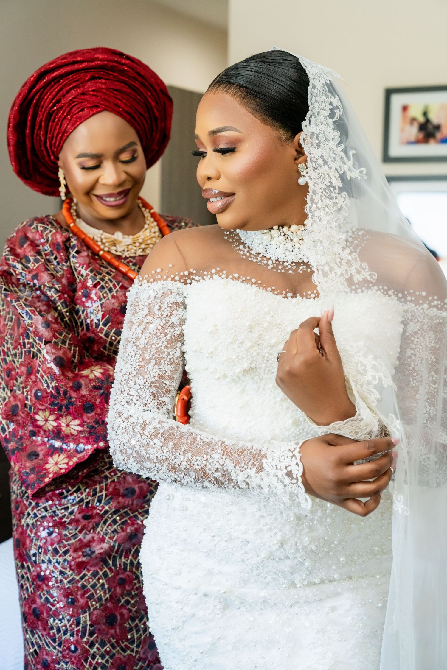 Intimate wedding preparation moment with bride in lace dress and guest in vibrant red traditional headwrap.
